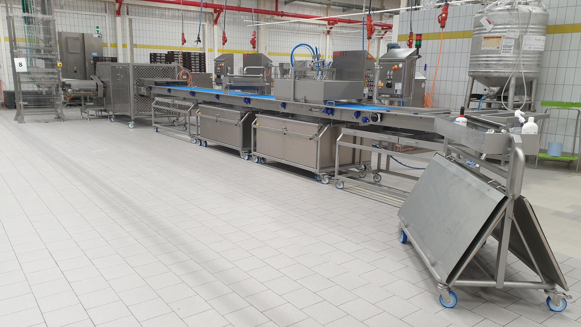 Eine moderne Lebensmittelverarbeitungsmaschine mit Förderbändern und Geräten aus rostfreiem Stahl in einer sauberen, industriellen Fabrikumgebung, umgeben von gefliesten Böden und Wänden.
