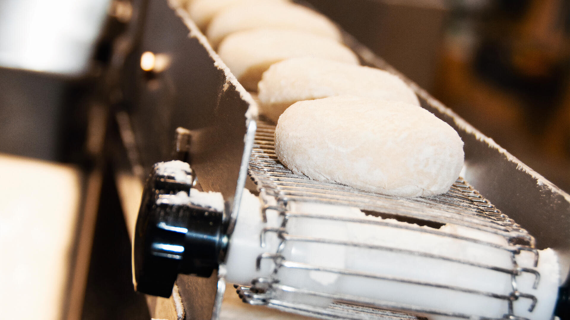 Close-up of several dough balls covered in flour moving along a metal conveyor belt in a bakery or food processing facility.