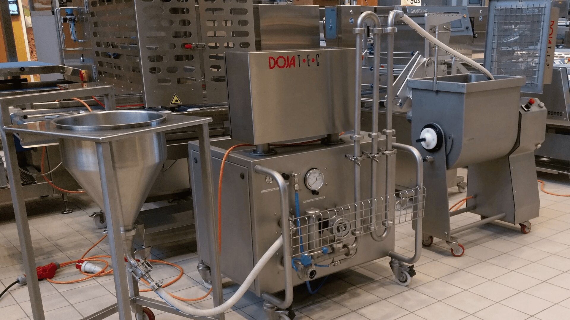 Industrielle Lebensmittelverarbeitungsanlagen mit Tanks, Rohren, Messgeräten und einem großen Mischer aus Edelstahl, die auf einem gefliesten Boden in einer Fabrikumgebung angeordnet sind. Orangefarbene Stromkabel verbinden die Maschinen miteinander.