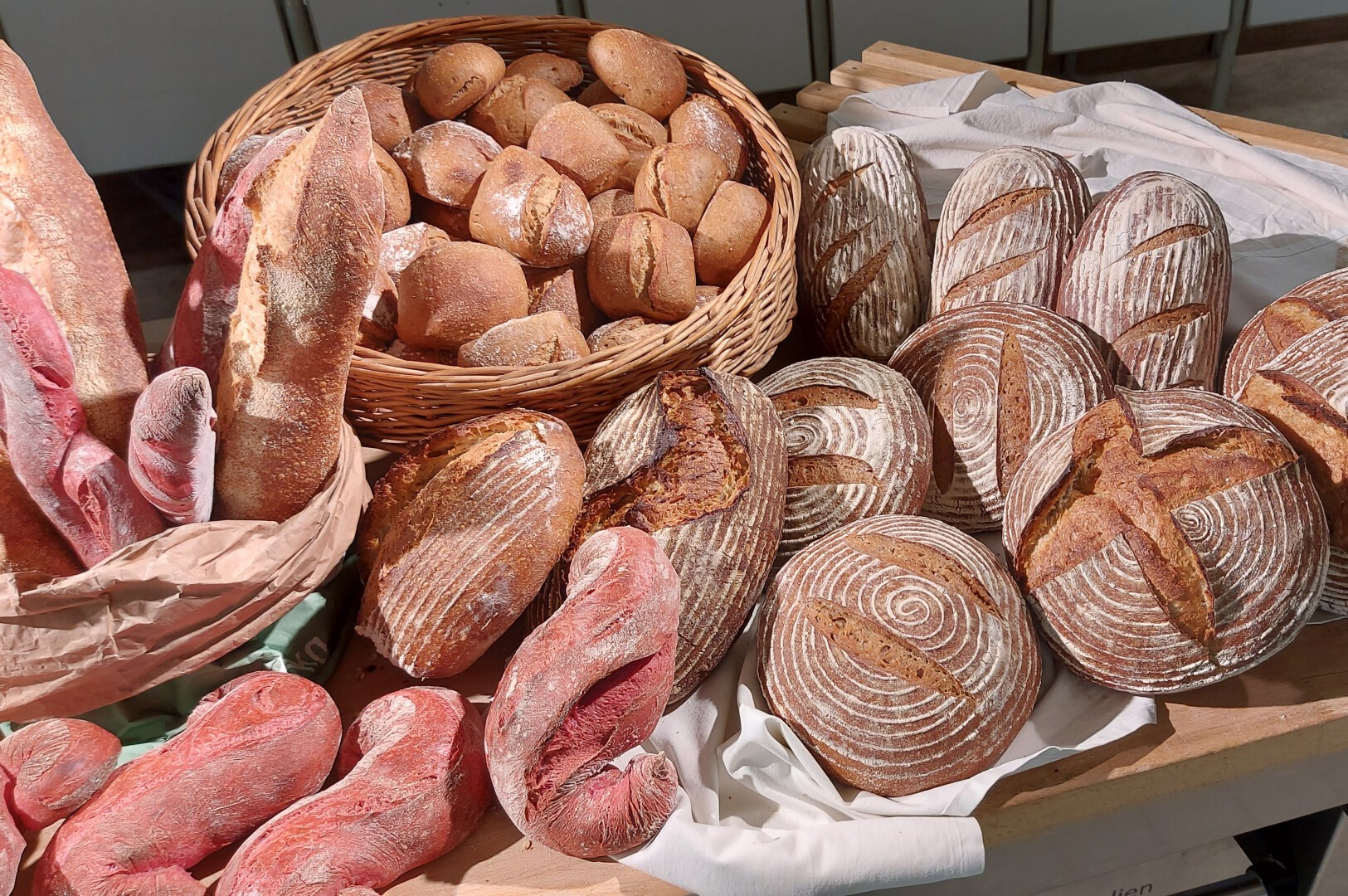 Eine Auswahl verschiedener rustikaler Brote, darunter runde Brote mit dekorativer Kruste, ovale Brote, rot getönte Baguettes und ein Korb mit kleinen runden Brötchen. Die Brote sind auf einem Holztisch angeordnet.