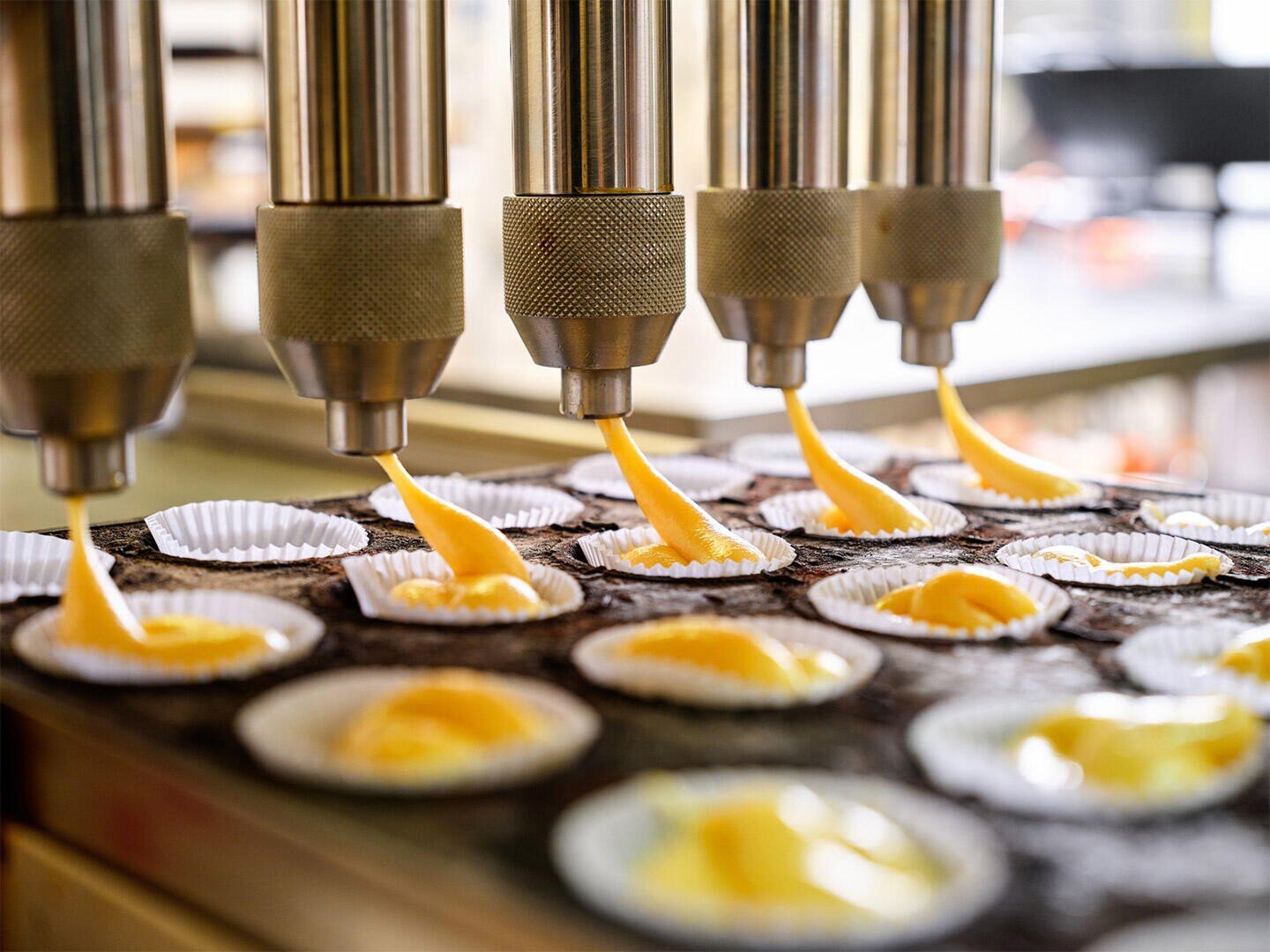 An einer Produktionslinie in einer Bäckerei oder Fabrik geben mehrere Düsen automatisch gelben Teig in Cupcake-Förmchen ab und bereiten so Cupcakes oder Muffins zum Backen vor.