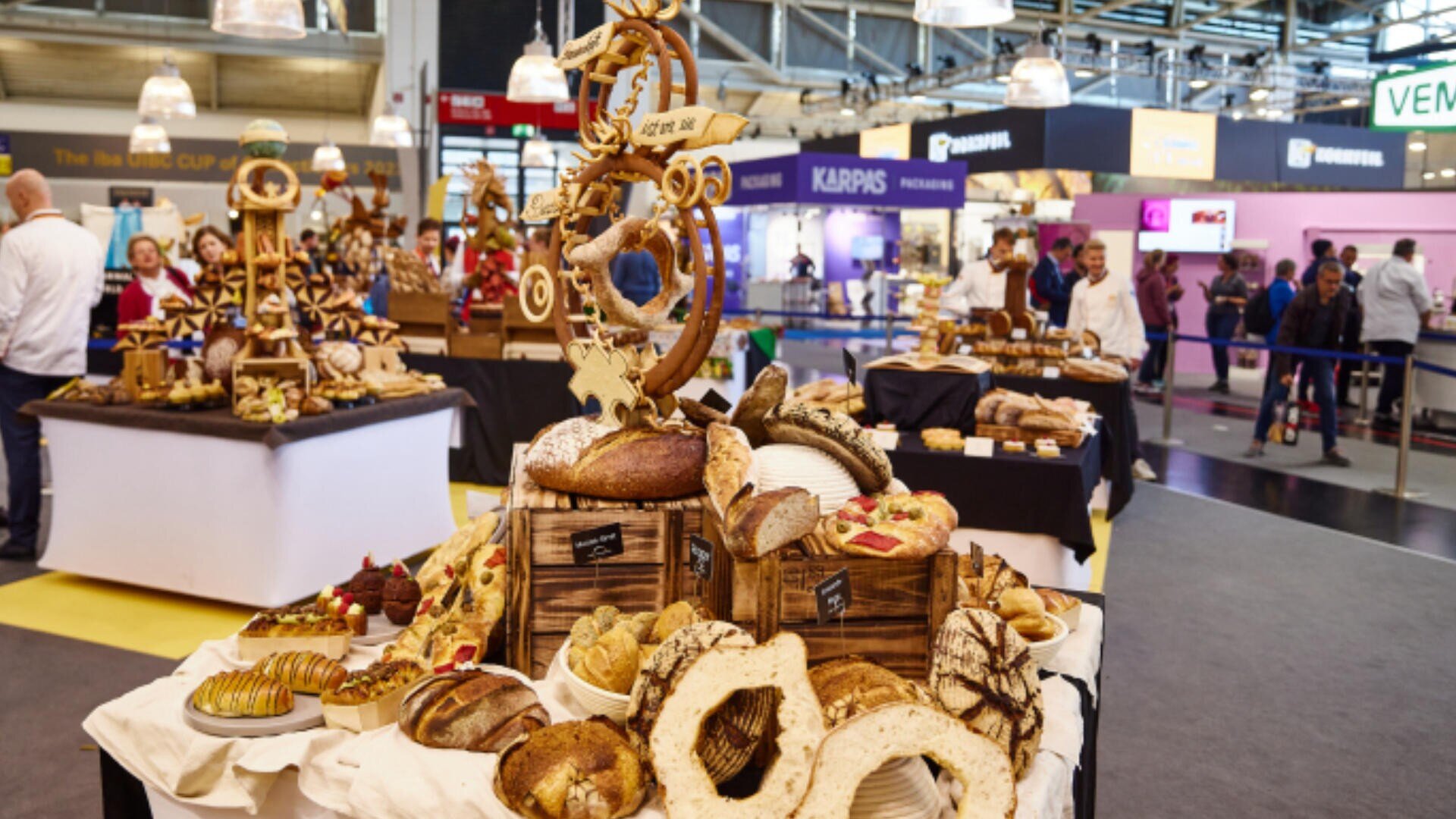 Ein Tisch auf einer Lebensmittelausstellung zeigt eine Auswahl an handwerklich hergestelltem Brot und Gebäck, mit einer großen dekorativen Brotskulptur in der Mitte. Im Hintergrund sind Menschen und Stände zu sehen.