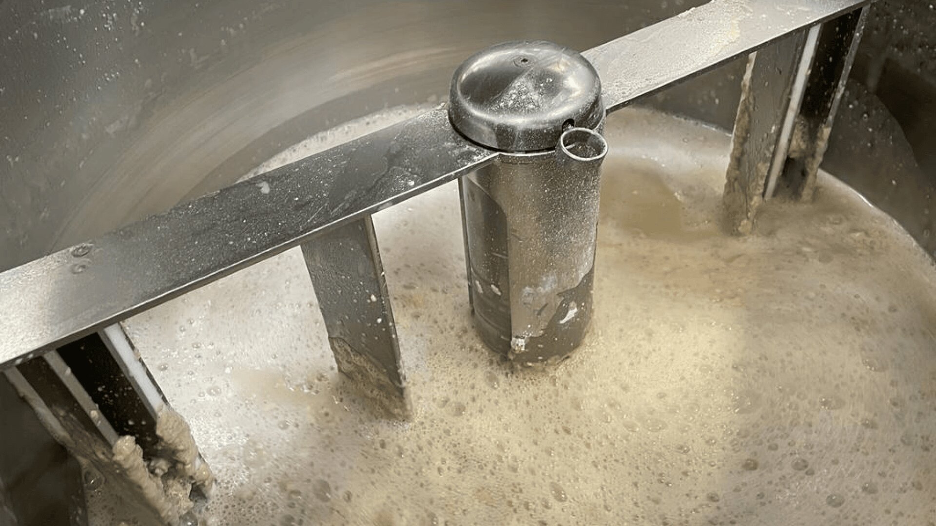 Close-up of an industrial mixer blending a foamy, beige liquid, likely in a metal container for food or beverage production. Metal paddles stir the mixture, creating bubbles and small lumps.