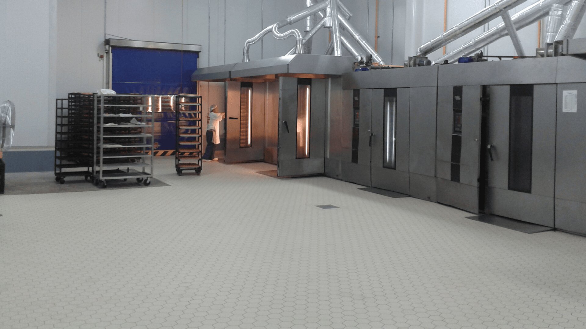 A commercial bakery with industrial ovens, metal racks holding baking trays, and a person in a white coat operating equipment. The floor is tiled and large ventilation ducts run along the ceiling.
