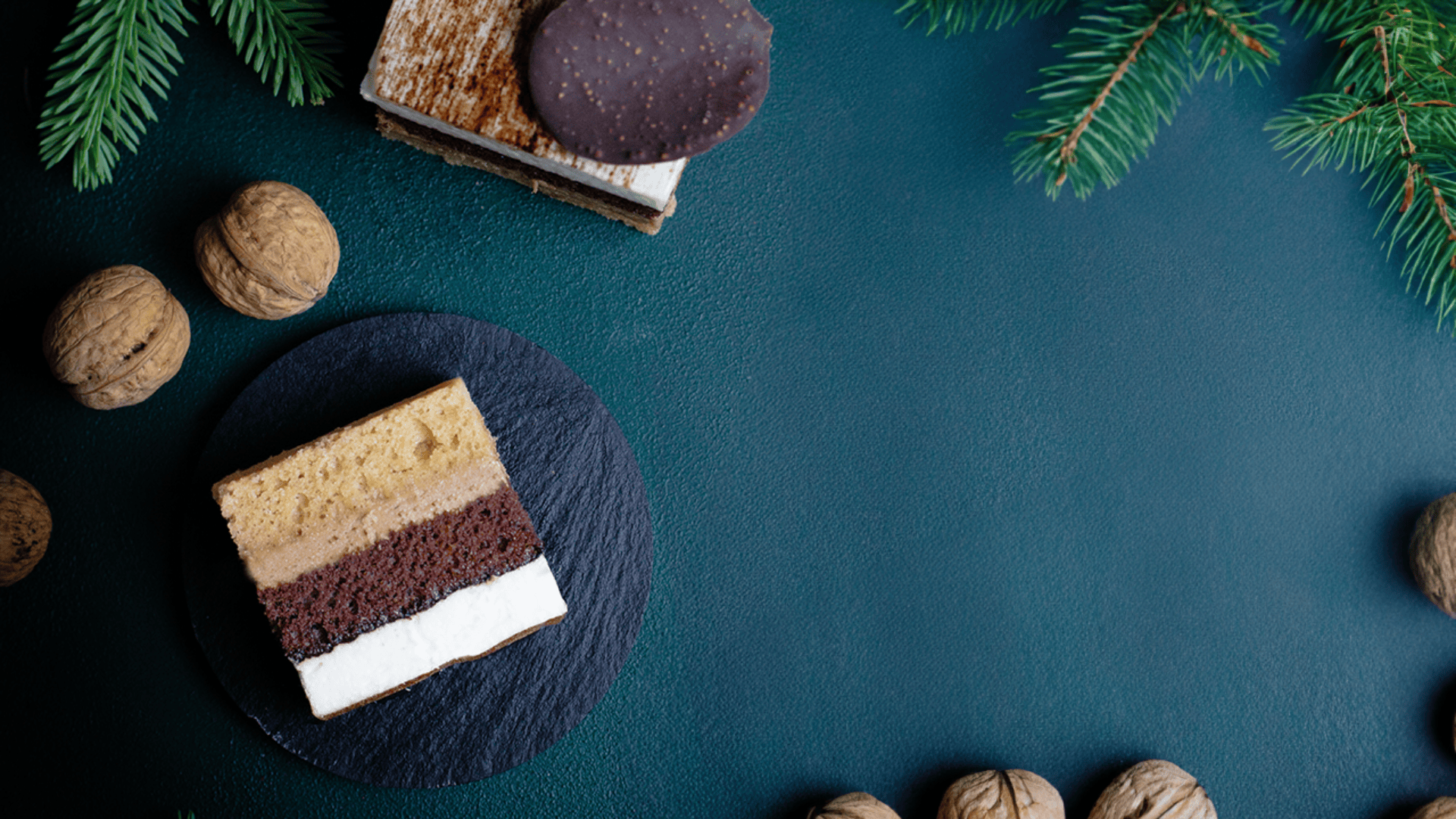 A piece of layered cake sits on a dark round slate, surrounded by whole walnuts and fir tree branches on a dark green surface. Another piece of cake with a chocolate disk is in the upper left corner.