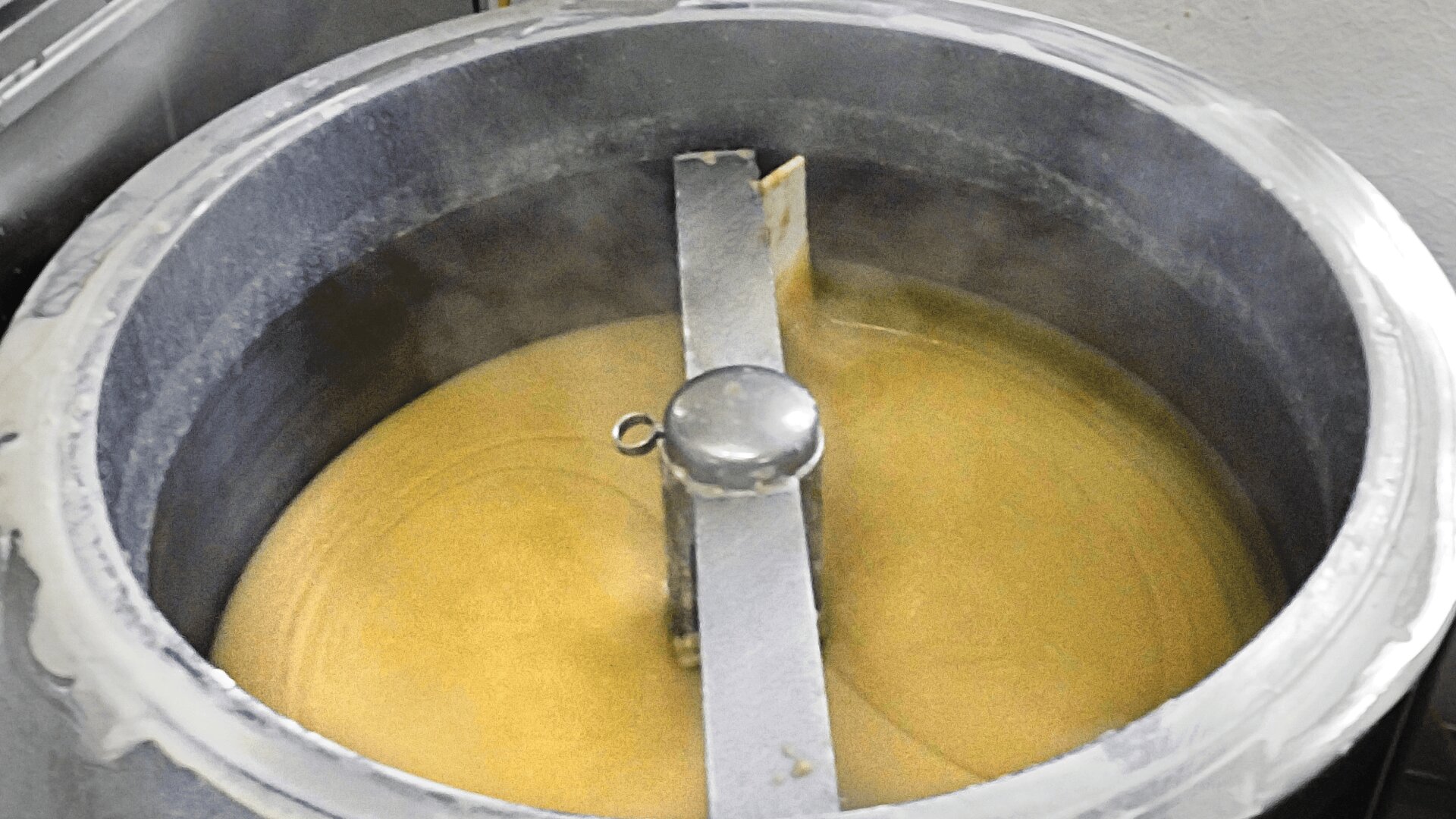Ein großer industrieller Metallmixer rührt eine dicke, hellbraune Flüssigkeit oder einen Teig. Aus der Mischung steigt Dampf auf, was bedeutet, dass sie heiß ist und verarbeitet wird.