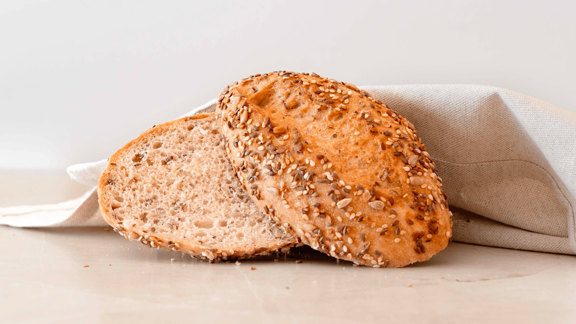 Two slices of multigrain bread rest on a light surface, partially covered by a beige cloth, against a plain light background. The bread is topped with assorted seeds.