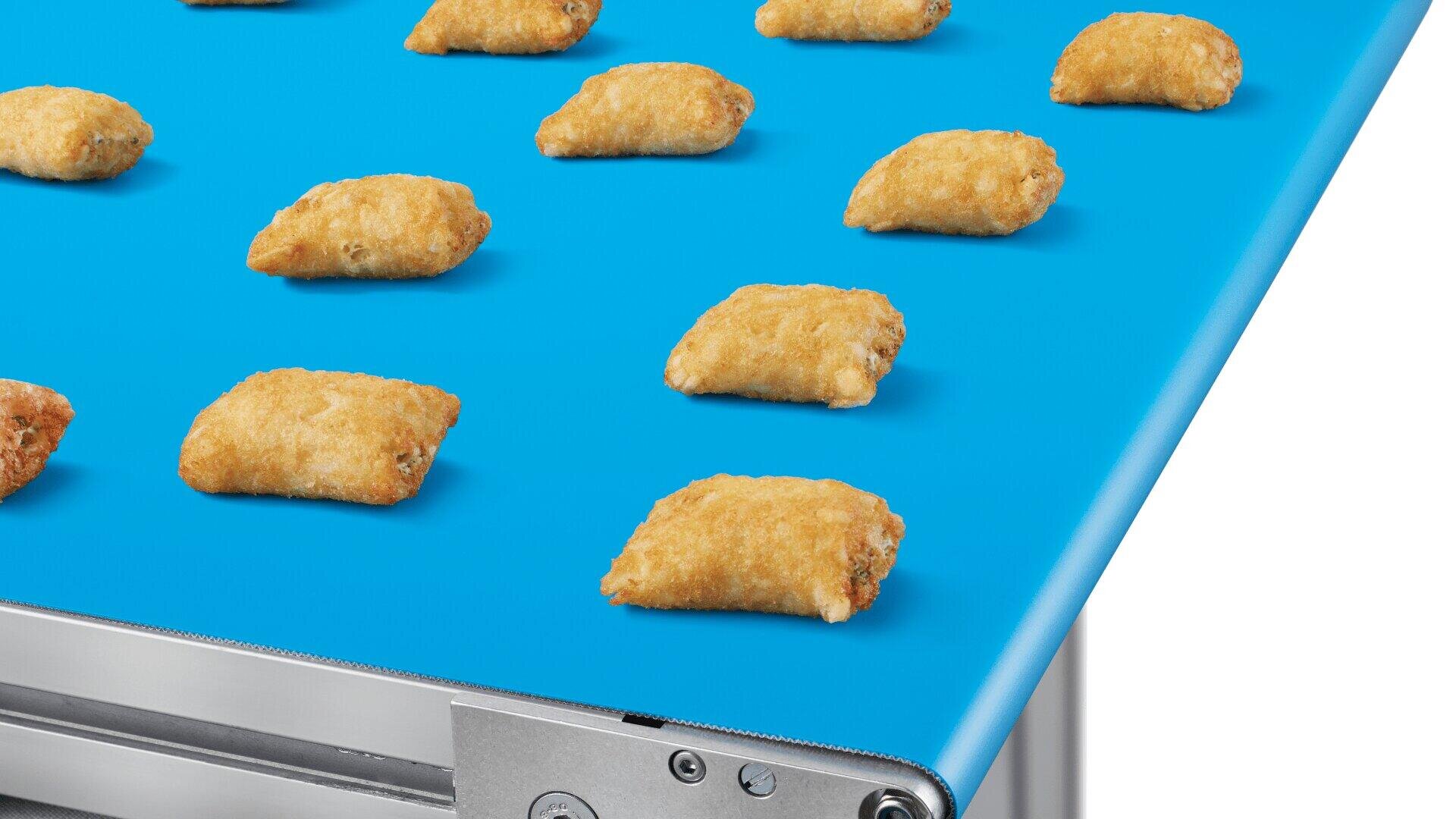 Small, golden-brown, breaded snack bites are evenly spaced on a bright blue industrial conveyor belt, likely in a food production facility.
