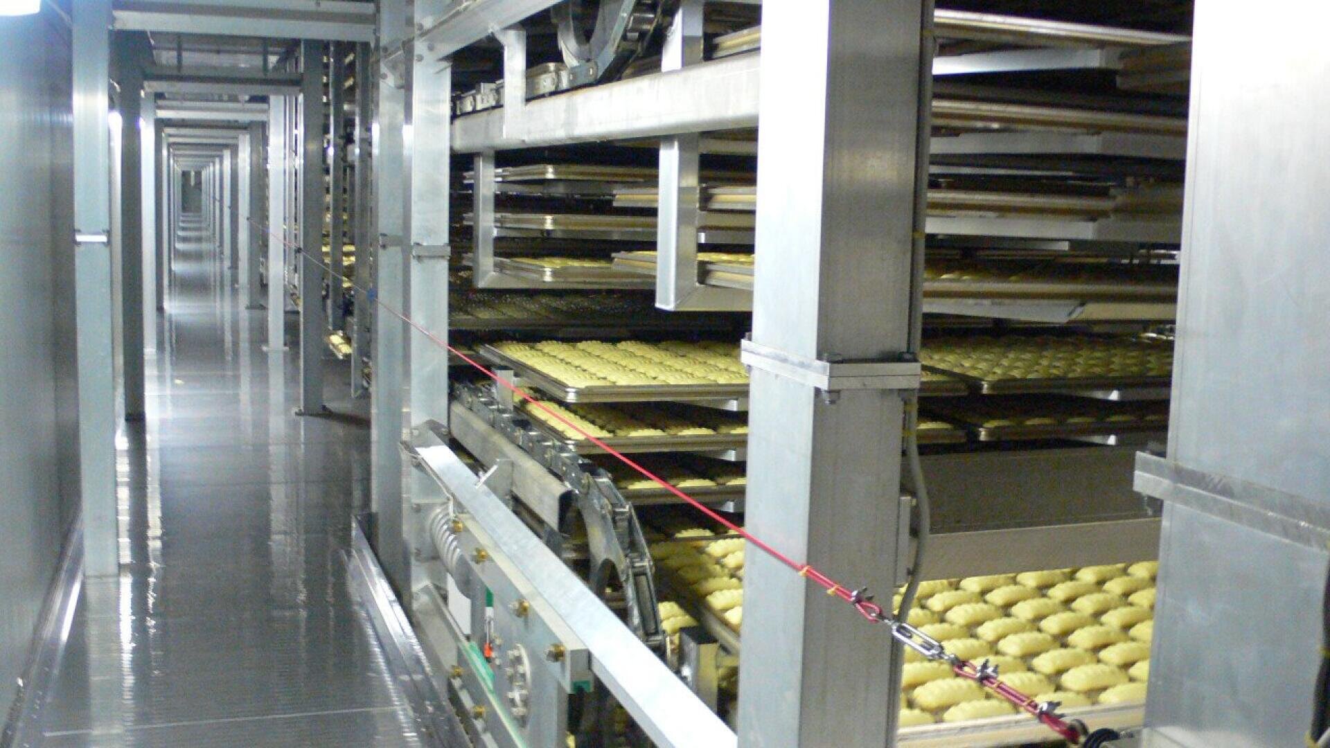 Reihen von Brotteig auf Blechen bewegen sich durch eine große industrielle Backmaschine in einer Fabrikumgebung mit Metallgestellen und einem Laufsteg entlang der Anlage.