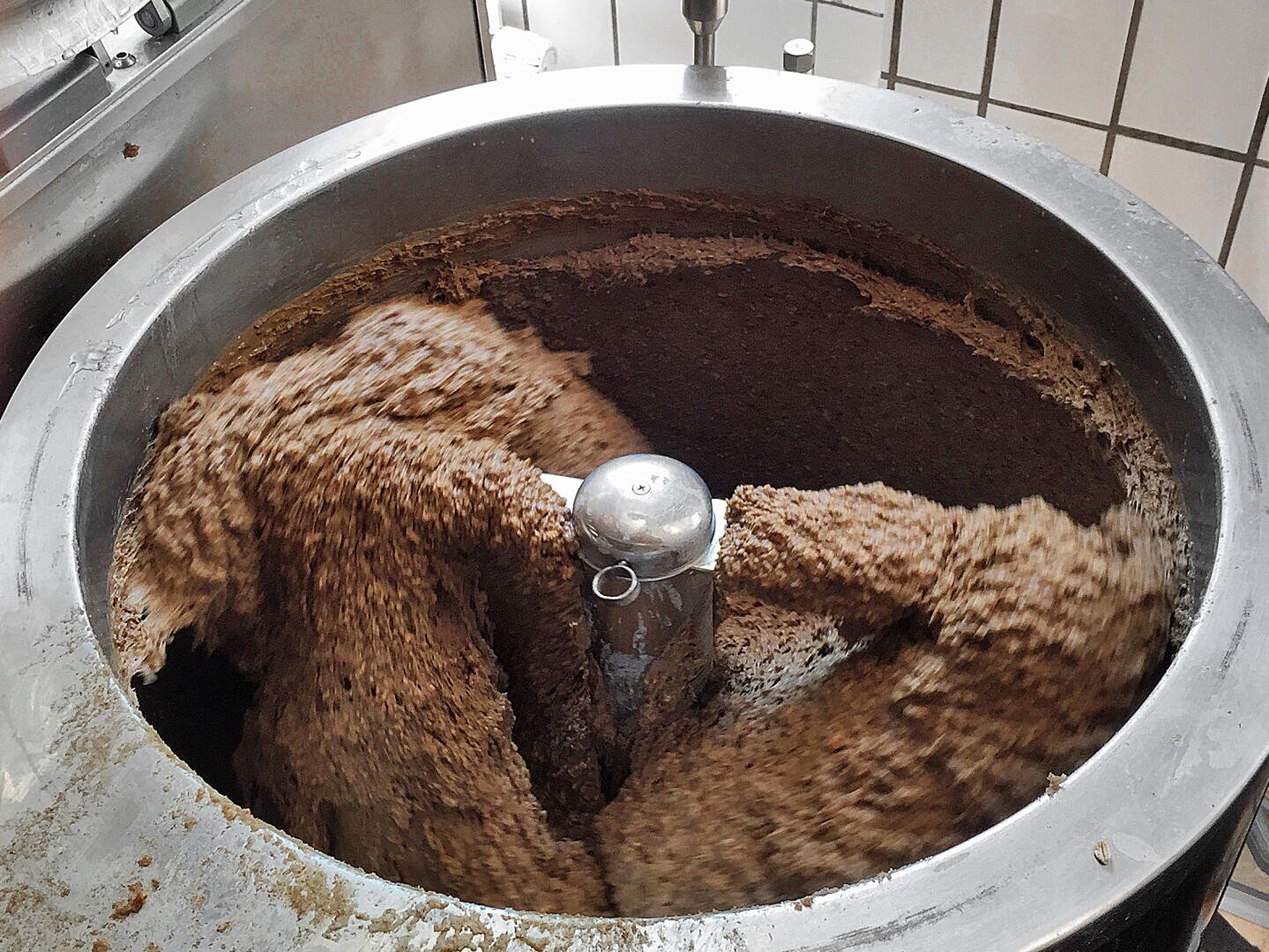 A large industrial mixer blends a thick, brown mixture inside a metal container, with the contents swirling around the central mixing blade. White tiled walls are visible in the background.
