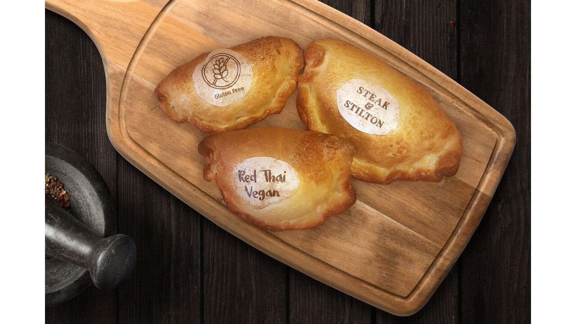 Three baked pasties with labels “Gluten Free,” “Steak & Stilton,” and “Red Thai Vegan” sit on a wooden cutting board atop a dark wooden table, next to a metal mortar and pestle.