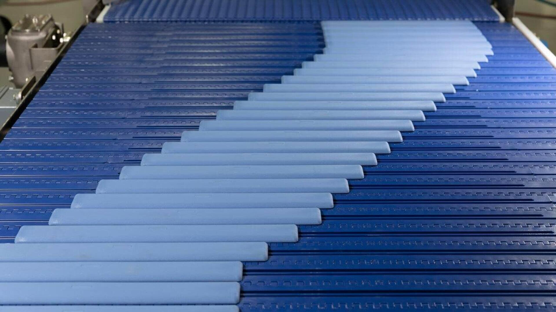 Hellblaue, rechteckige Objekte bewegen sich diagonal auf einem dunkelblauen Förderband in einer Fabrik oder einem Produktionsbetrieb und bilden ein Zickzackmuster auf der Oberfläche.