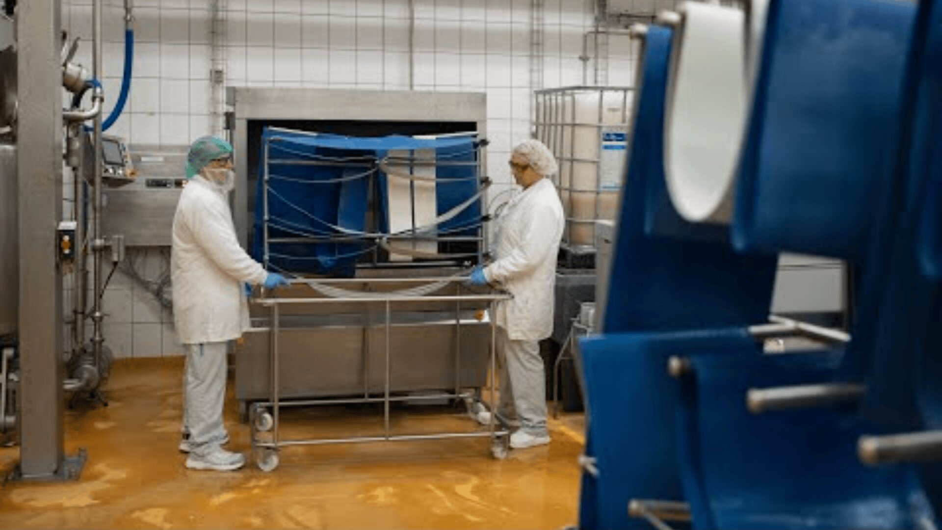 Zwei Arbeiter in weißen Laborkitteln, mit Haartüchern und Masken hantieren mit Geräten in einer sauberen, industriellen Lebensmittelverarbeitungsanlage mit gekachelten Wänden und gelbem Boden. Im Vordergrund sind blaue Maschinen zu sehen.