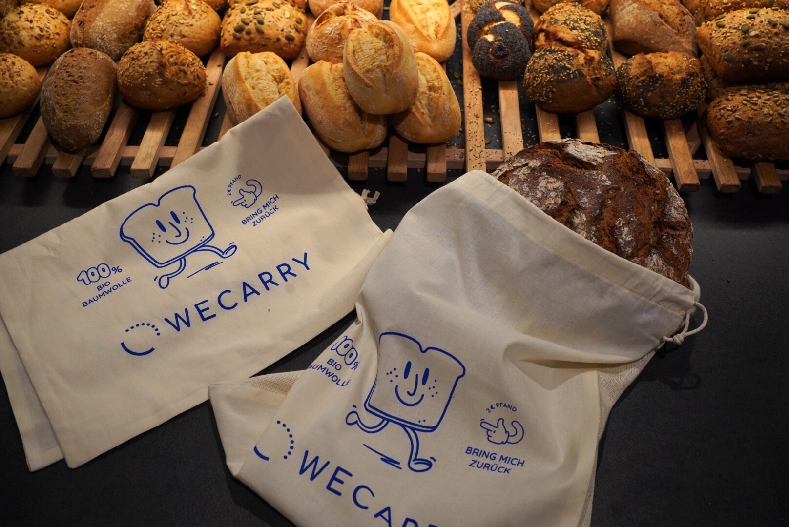 Wiederverwendbare Brotbeutel aus Stoff mit einem lächelnden Brotscheiben-Cartoon und dem Text „WeCarry“ werden auf einer Theke präsentiert, während im Hintergrund verschiedene frische Brötchen und Brote auf Holzregalen angeordnet sind.