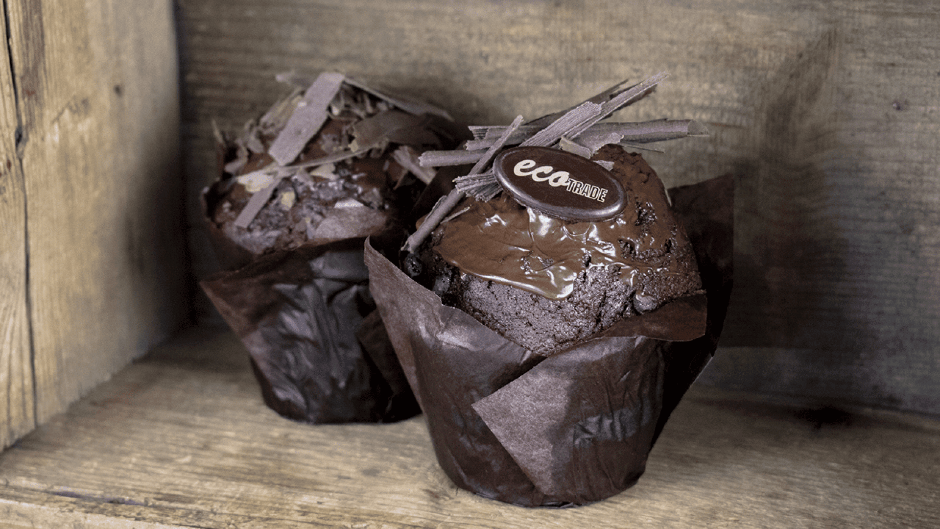 Zwei reichhaltige Schokoladenmuffins in braunen Verpackungen sind mit Schokoladenspänen und einer kleinen runden Schokolade mit der Aufschrift eca vor einem rustikalen Holzhintergrund versehen.