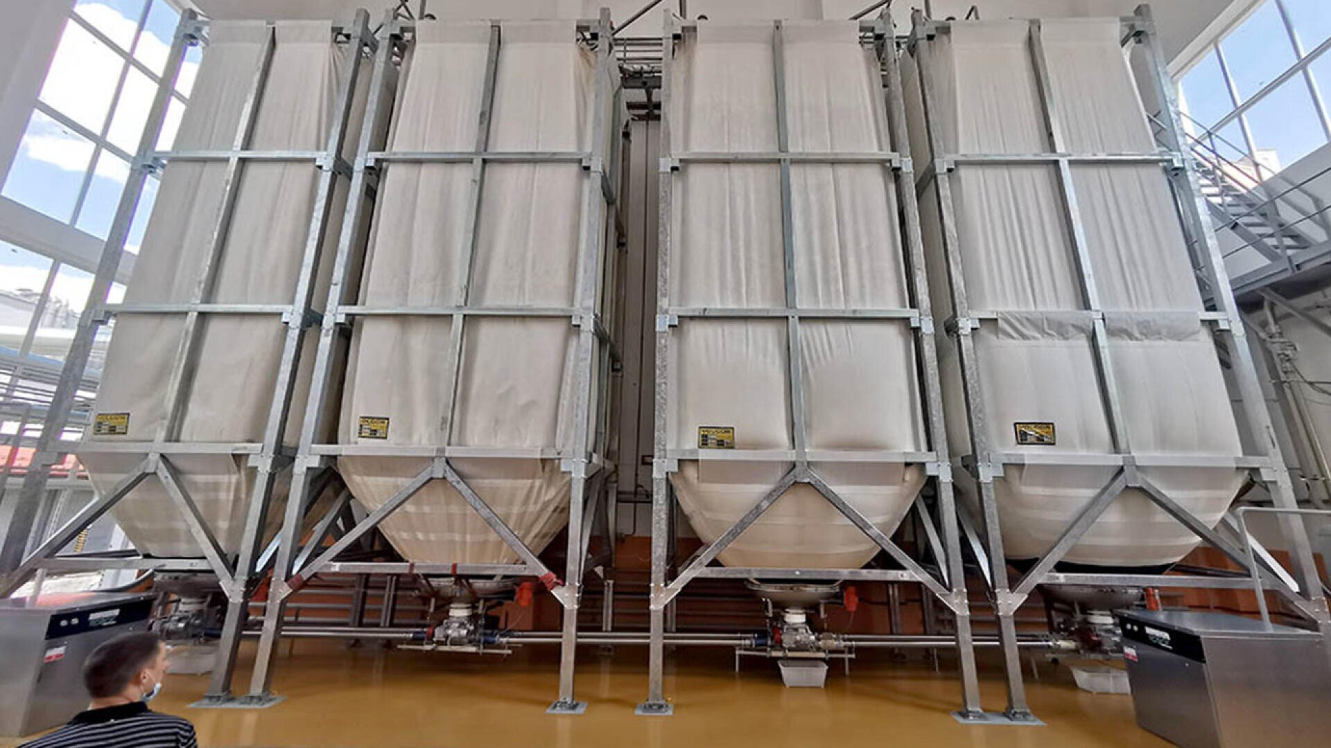 Große, von Metallrahmen getragene Industriesilos für Textilien stehen in einer hellen Anlage. Unten sind Maschinen und Rohre zu sehen und in der unteren linken Ecke ist eine Person zu sehen, die den Aufbau beobachtet.