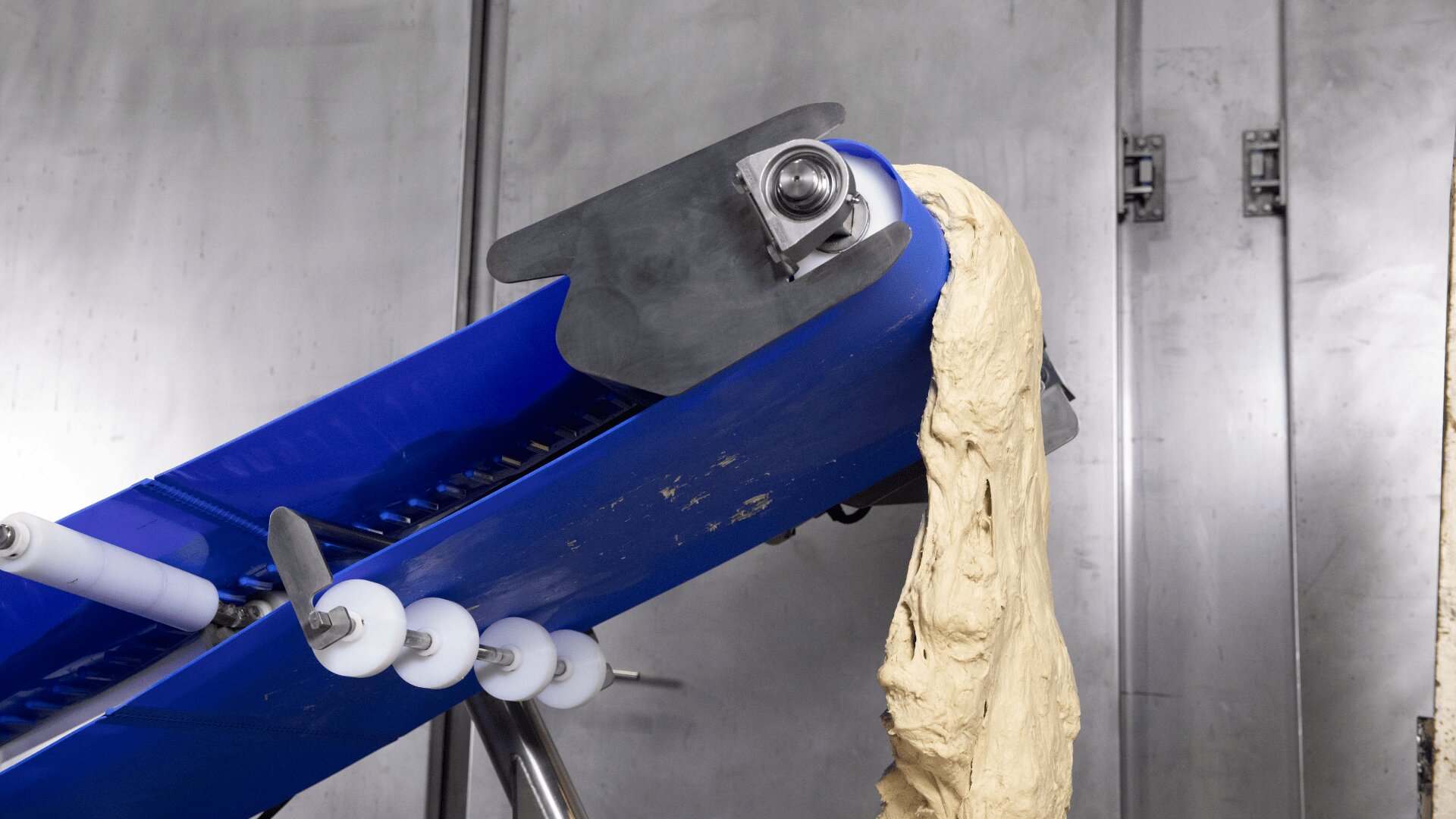 A blue conveyor belt carries raw dough upwards in a food processing facility, with the dough hanging over the edge near metal machinery and a stainless steel background.