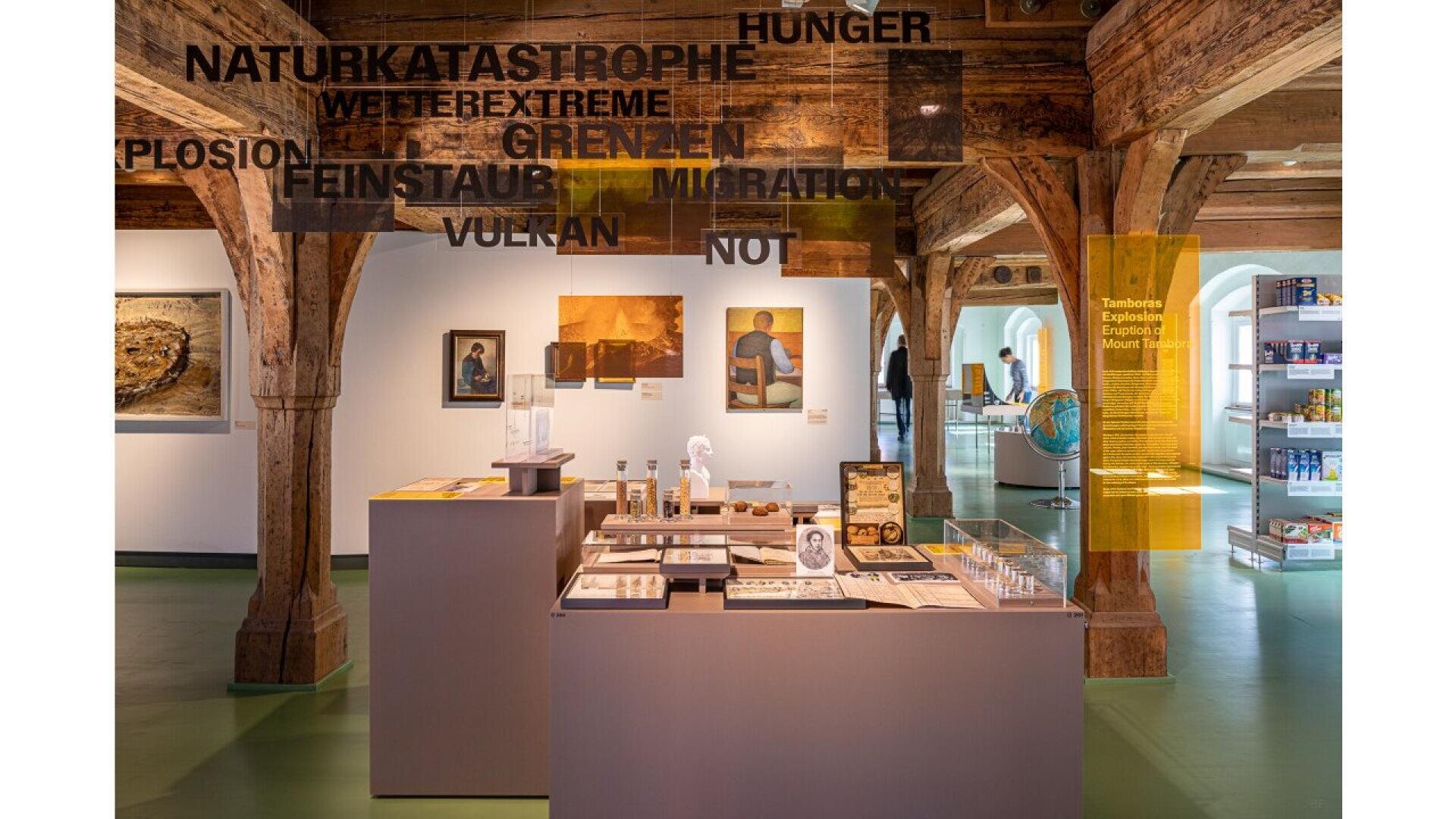Museum exhibit with wooden beams, display tables featuring various artifacts, and large transparent panels overhead with German words like NATURKATASTROPHE and MIGRATION. Paintings and shelves are visible in the background.