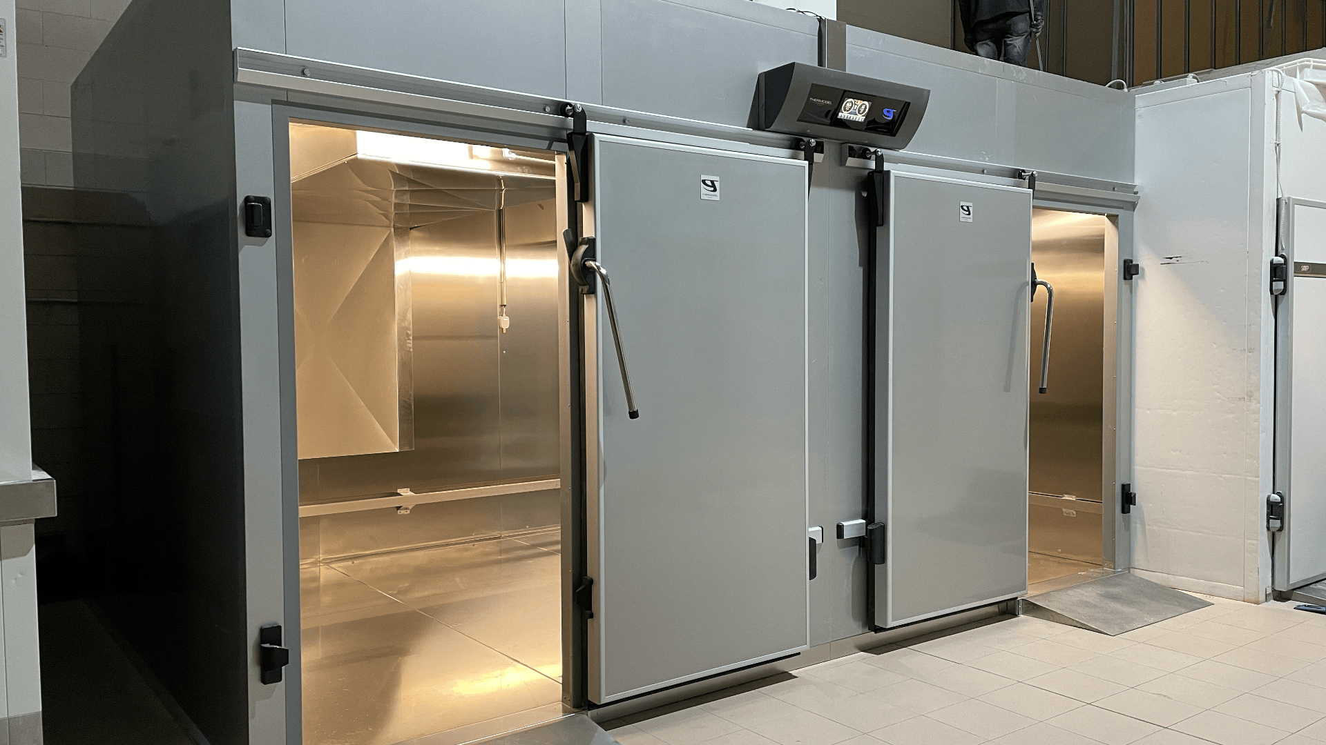 Two large industrial metal cold storage rooms with open doors reveal empty, clean interiors. Digital control panels are mounted above the doors, and the space has tiled flooring.