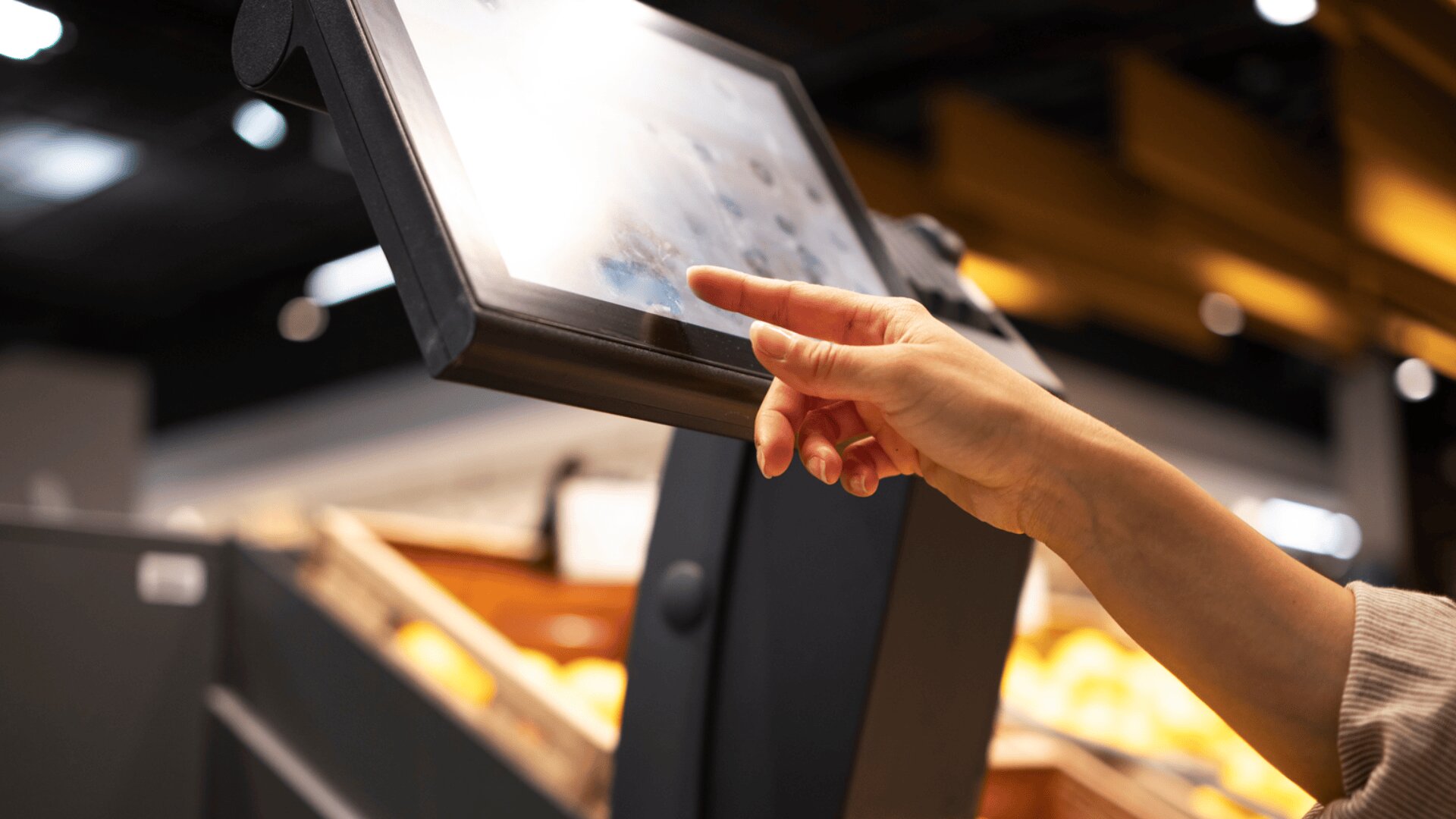 Eine Person berührt mit der Hand den Bildschirm eines Selbstbedienungskiosks in einem Geschäft, wahrscheinlich zum Bezahlen oder für Informationen. Der Hintergrund ist unscharf und zeigt die Regale und Produkte des Geschäfts.