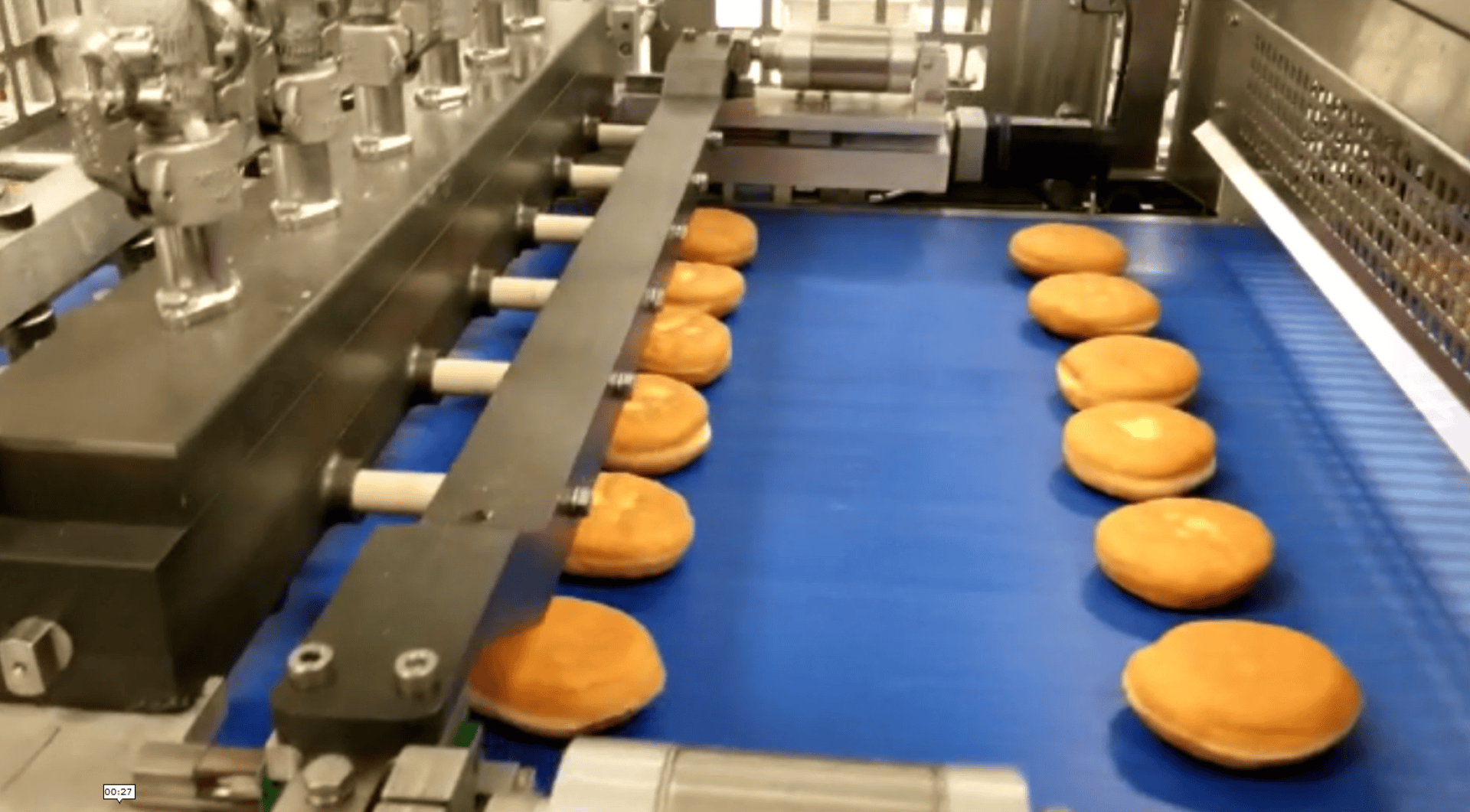 Rows of round doughnuts move along a blue conveyor belt in a factory, passing under machinery designed for mass production or filling.