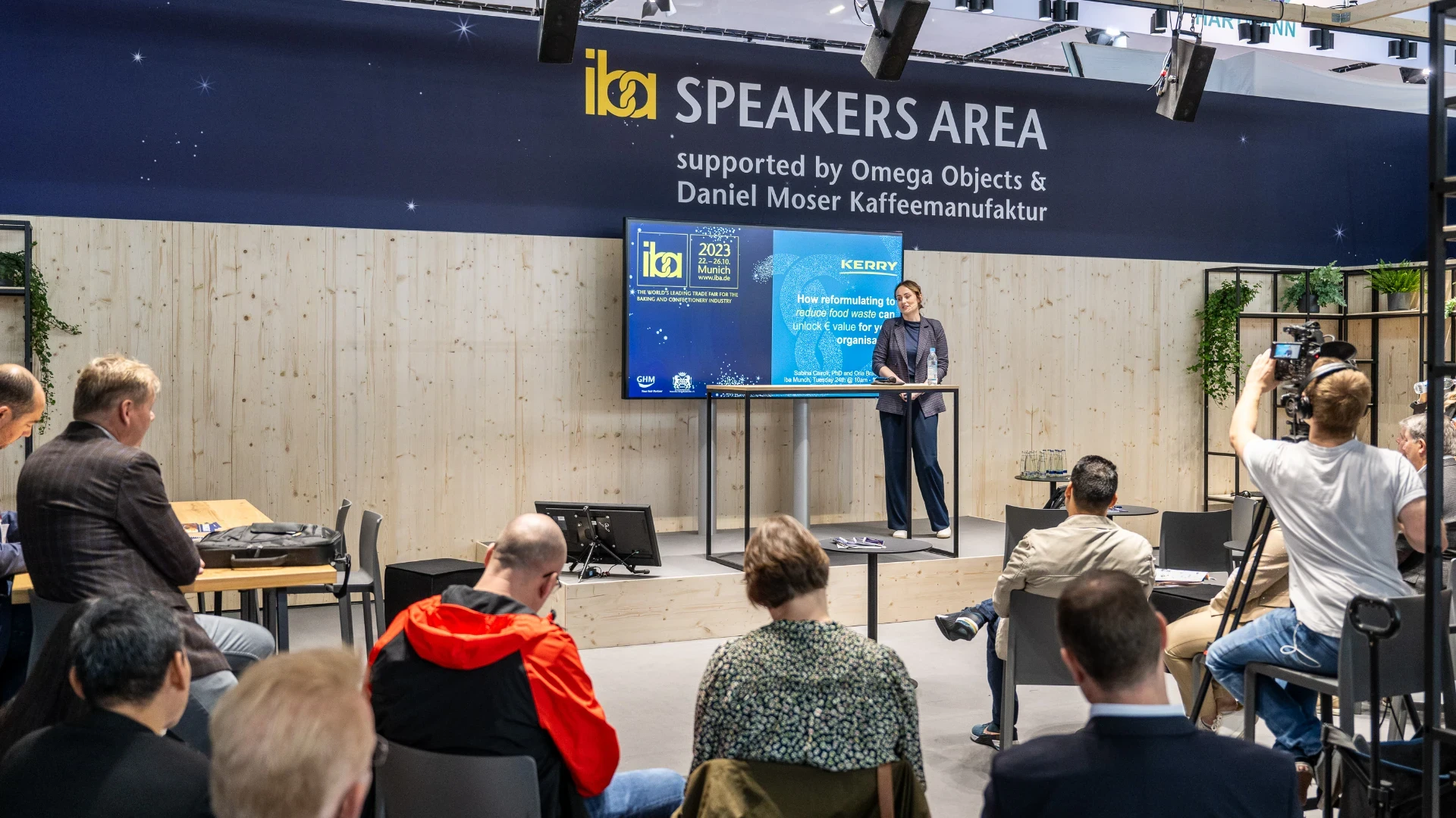 A speaker stands at a podium giving a presentation to an audience in the SPEAKERS AREA at a conference. Attendees are seated and listening, some taking notes, while others photograph or film the event.