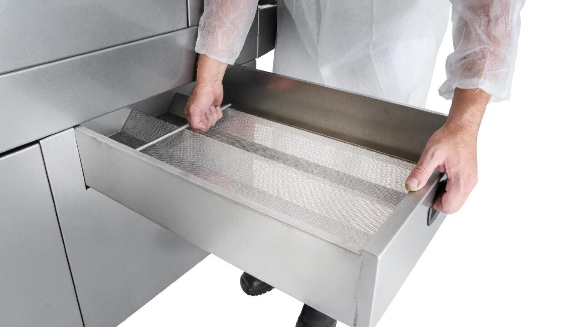 A person wearing a white lab coat and gloves pulls out a metal drawer with mesh trays from a stainless steel industrial machine.