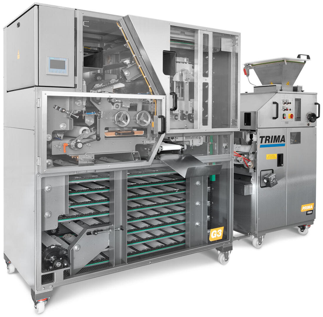 Eine große industrielle Brotschneide- und Verpackungsmaschine aus Edelstahl, mit sichtbaren Zahnrädern und Riemen, mit den Bezeichnungen "TRIMA" und "G3", für den gewerblichen Einsatz in Bäckereien.