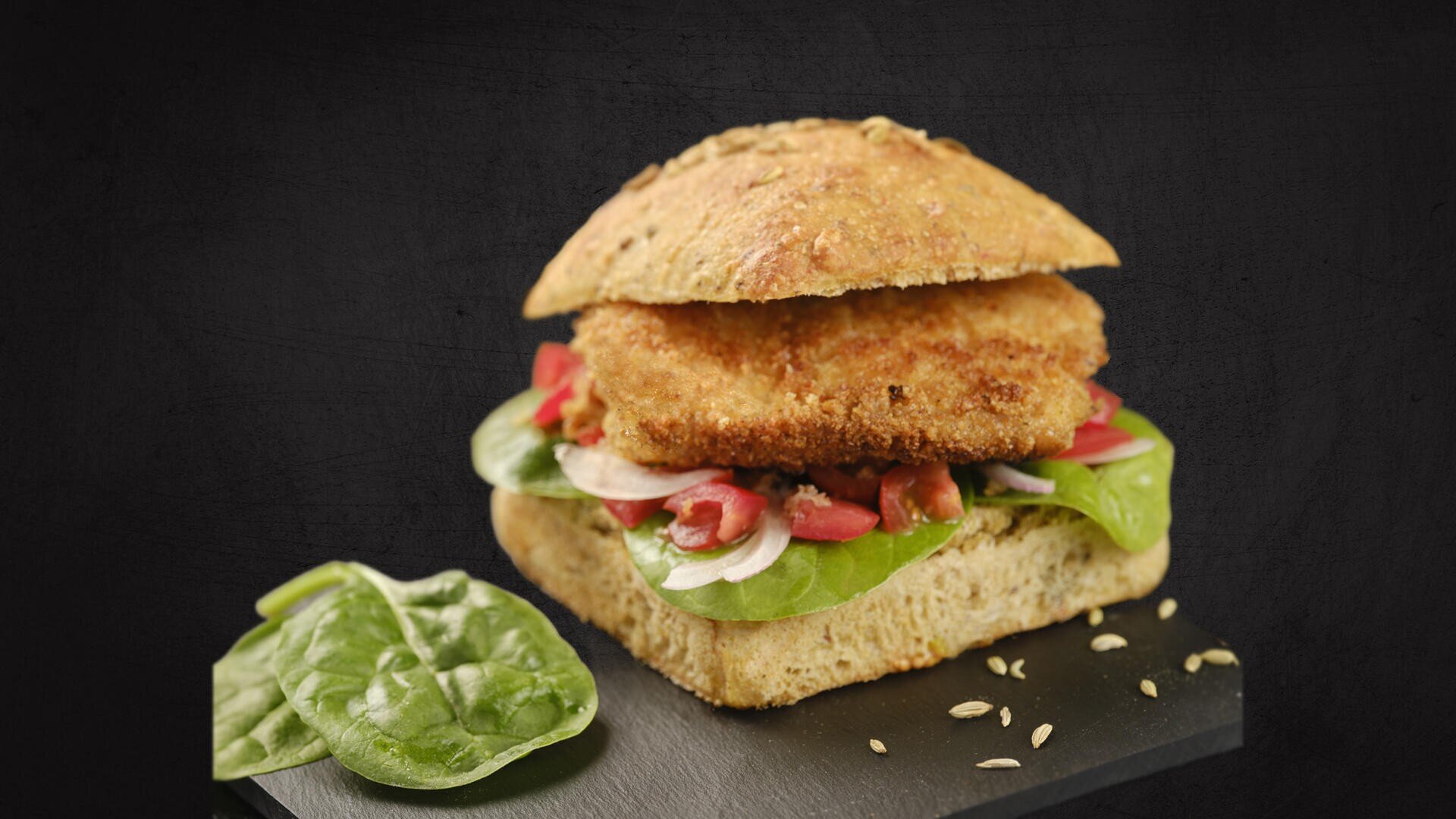 Ein Sandwich mit einem quadratischen Vollkornbrötchen, paniertem Patty, frischem Blattspinat, geschnittenen Tomaten und Zwiebeln. Ein Spinatblatt und Fenchelsamen liegen auf der Schiefertafel vor dem Sandwich.