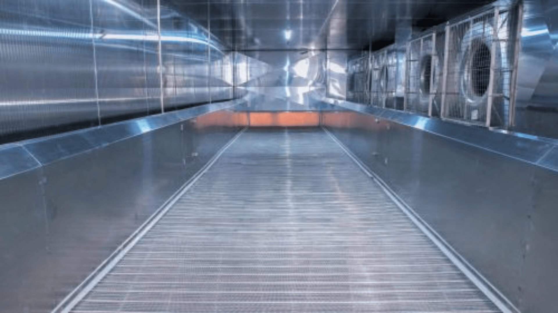 A wide, empty industrial corridor with metallic walls, grated flooring, and large circular ventilation fans on the right side, all surfaces reflecting light for a modern, sterile appearance.
