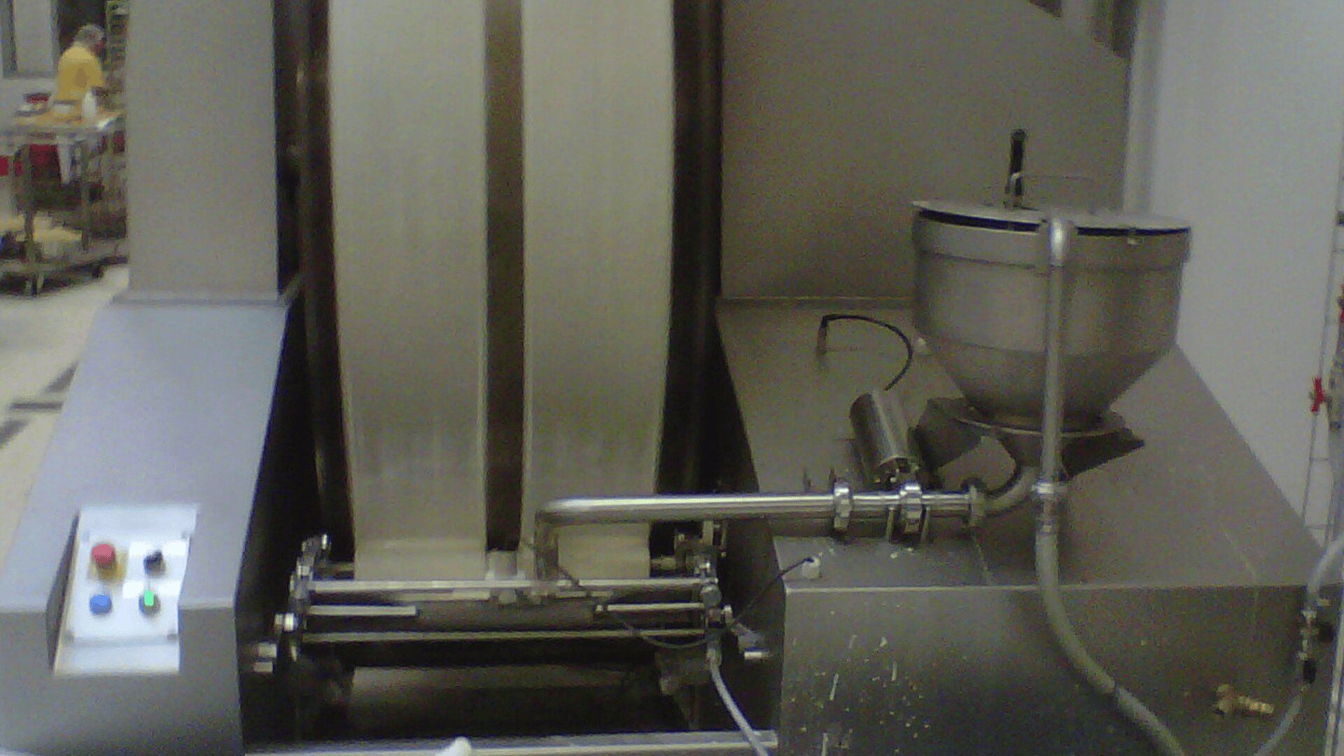 A close-up of industrial food processing equipment, featuring a metal bowl with attached pipes, a control panel with colored buttons, and a large vertical drum in a factory setting.