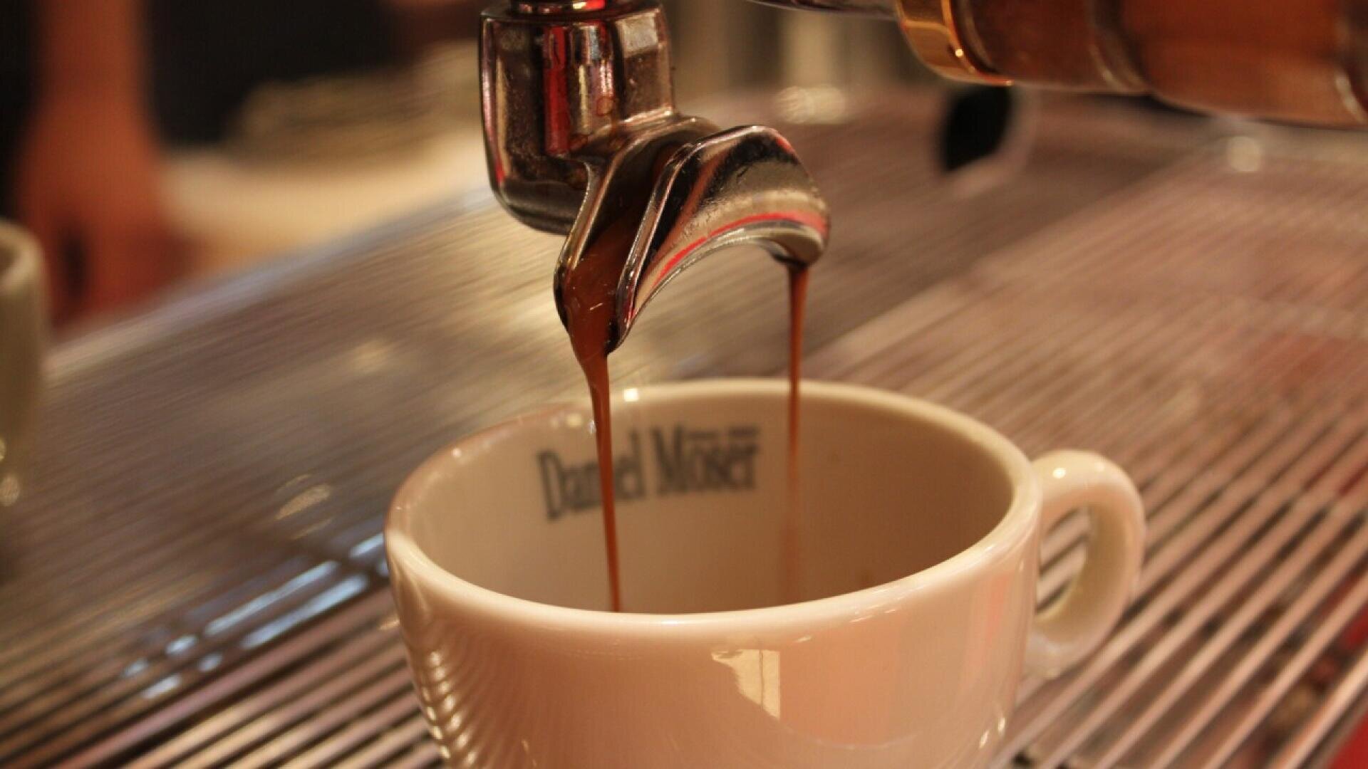 Close-up of an espresso machine pouring coffee into a white ceramic cup, with rich brown coffee streams flowing from the spout. The cup sits on a metal grate beneath the machine.