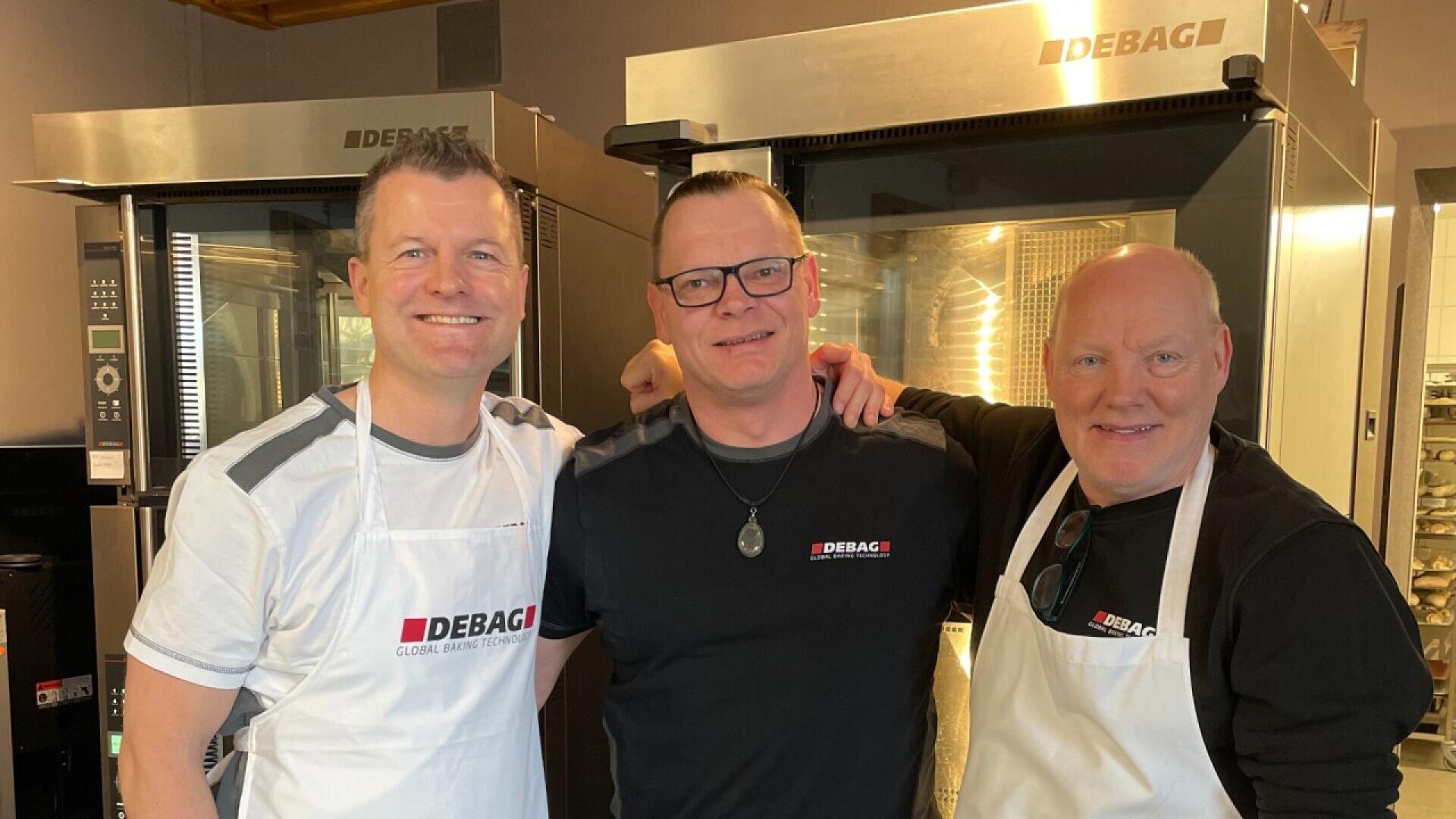 Three smiling men, two wearing white aprons and one in black, stand together in front of industrial ovens. All wear shirts with the DEBAG logo, suggesting they are in a commercial kitchen or bakery setting.