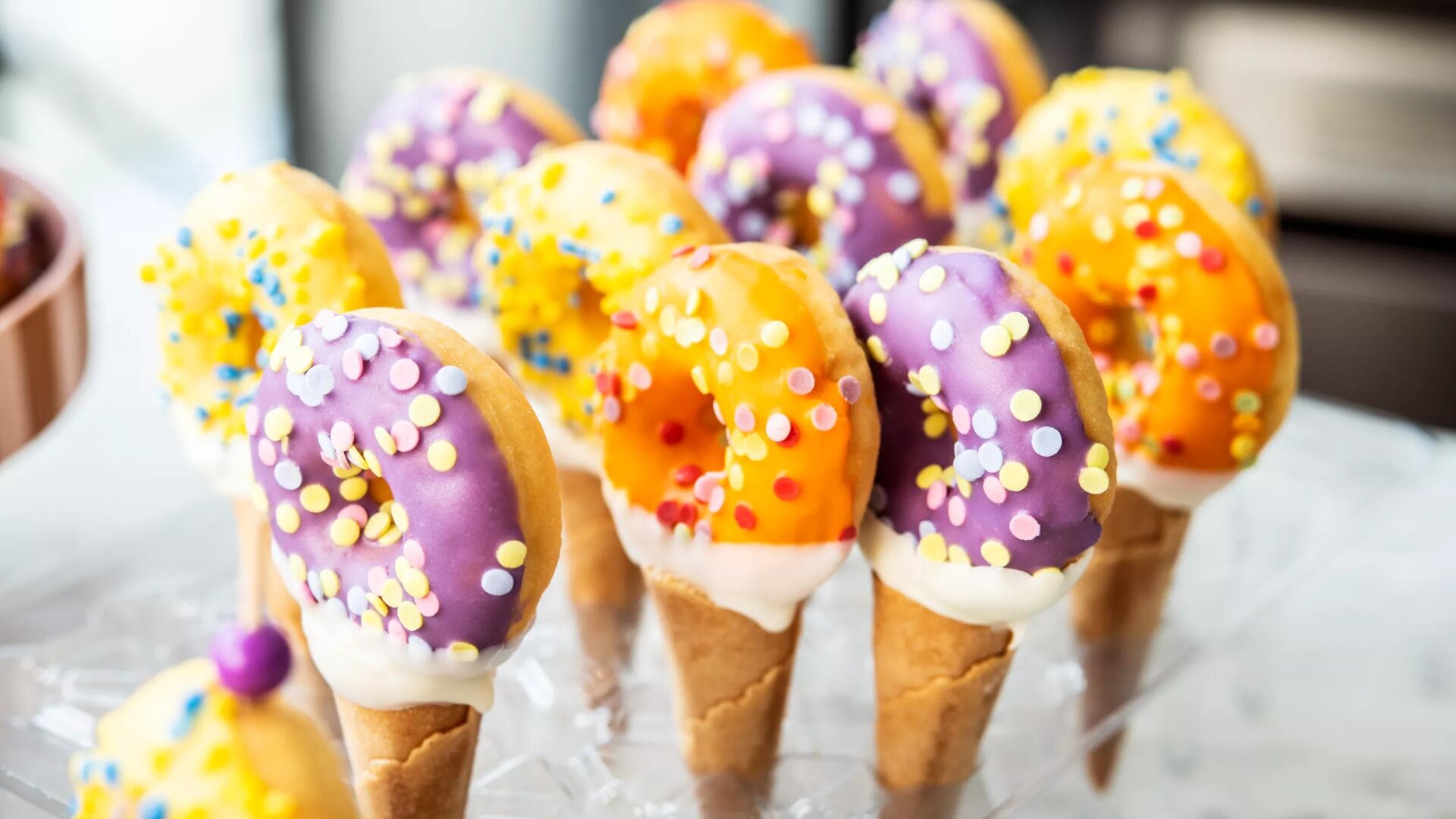 Bunte Donuts mit lilafarbenem und orangefarbenem Zuckerguss, die mit Streuseln bestreut sind, befinden sich auf mit weißem Zuckerguss gefüllten Eistüten, die auf einem durchsichtigen Ständer angeordnet sind.