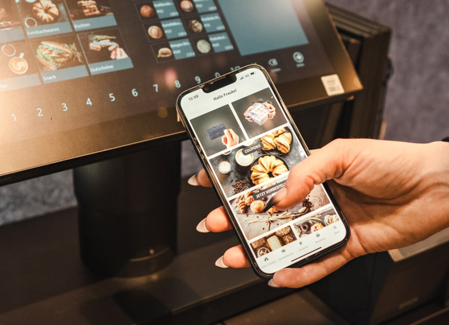 A person holds a smartphone displaying a food ordering app while standing in front of a digital touchscreen menu at a restaurant or café.