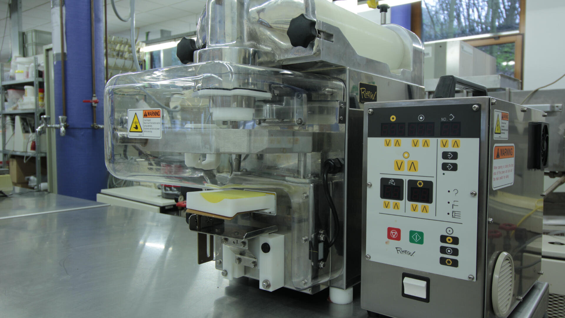 Eine Nahaufnahme einer industriellen Lebensmittelverarbeitungsmaschine mit Bedienfeld, Knöpfen, Warnschildern und einem Auffangbehälter für verarbeitete Lebensmittel in einer Großküche.