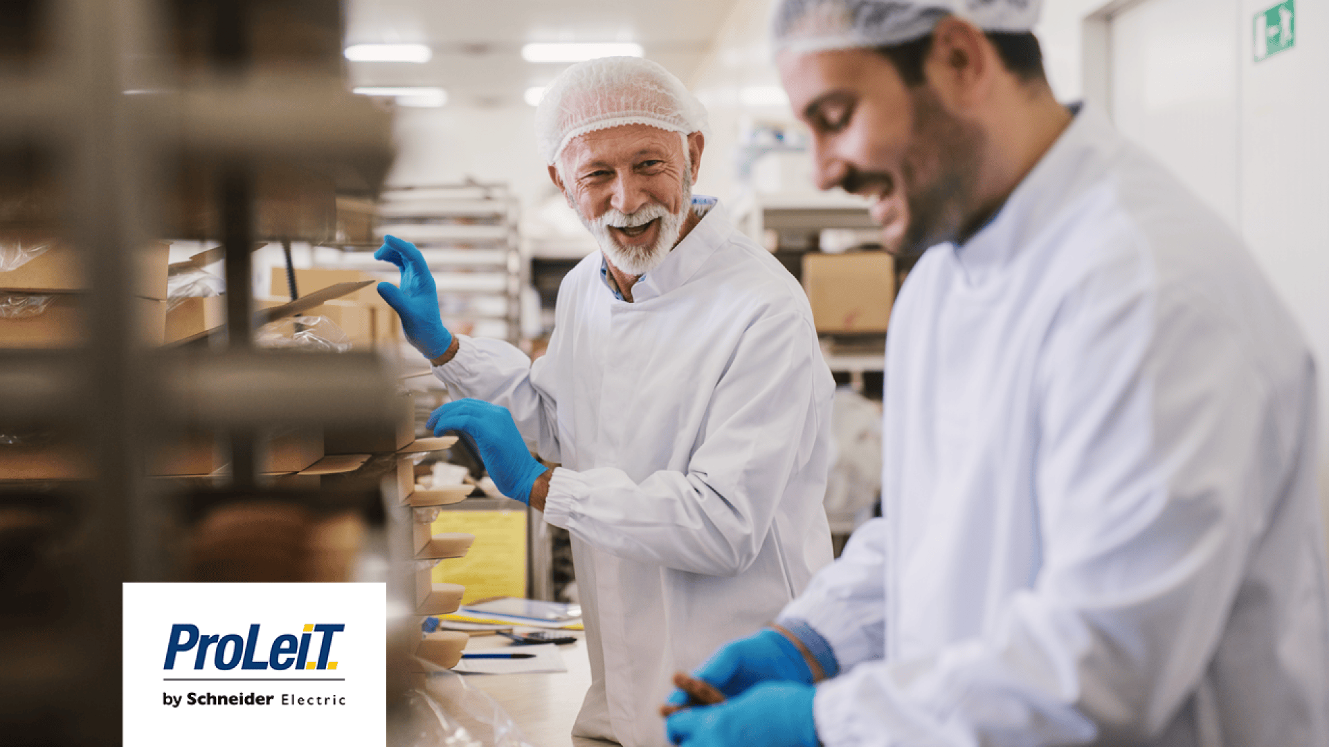 Zwei Männer in weißen Laborkitteln, Haarnetzen und blauen Handschuhen arbeiten zusammen in einer sauberen Lebensmittelproduktionsanlage. Sie lächeln und hantieren mit verpackten Waren. In der Ecke ist das Logo von ProLeiT by Schneider Electric zu sehen.