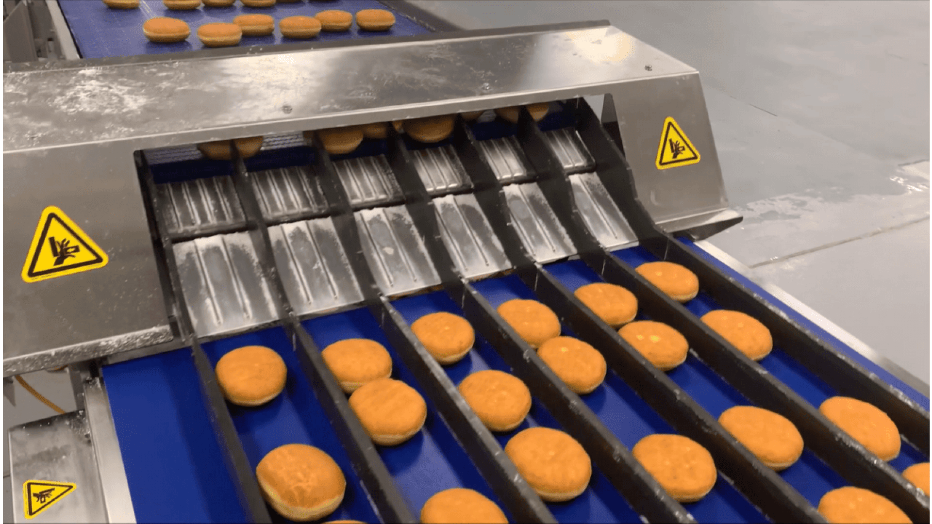 Reihen von runden Donuts bewegen sich über ein Förderband in einer Fabrik und laufen unter einer Metallmaschine mit Warnschildern, die auf eine elektrische Gefahr hinweisen. Der Boden und der umliegende Bereich wirken sauber und industriell.
