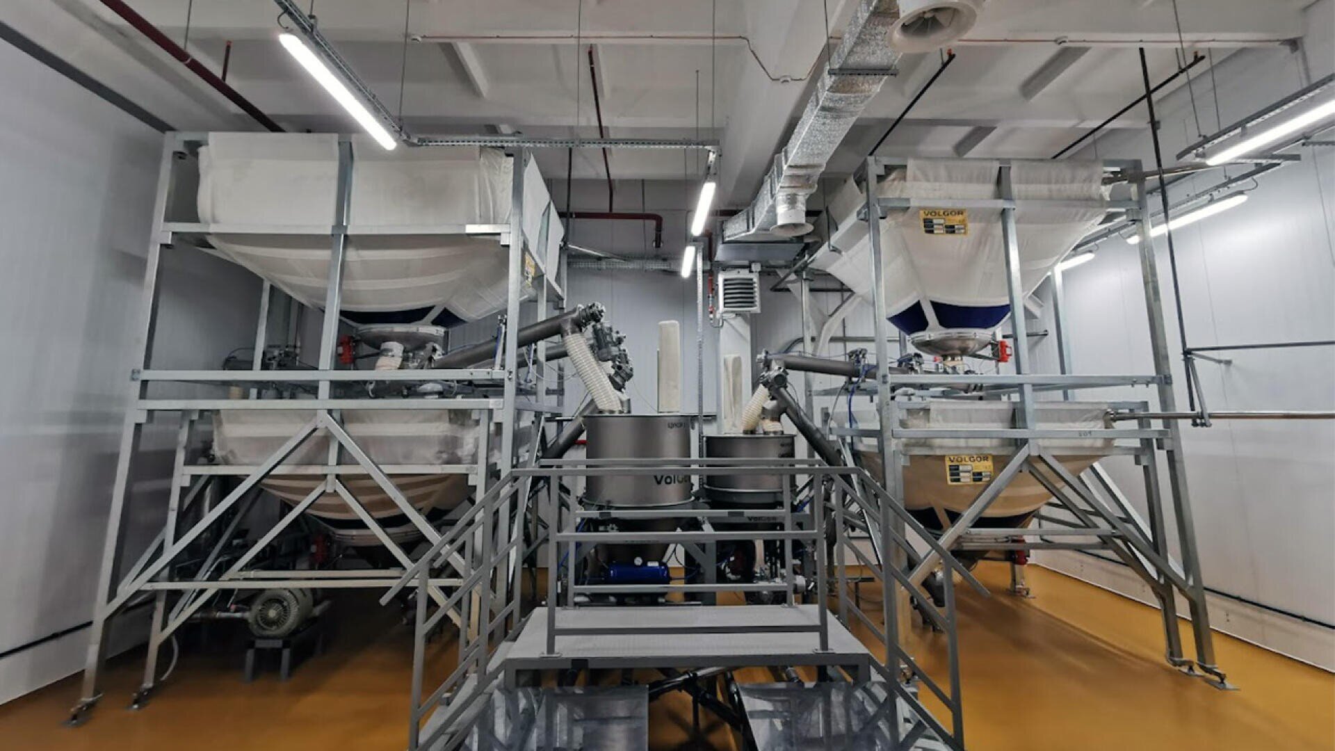 Zwei große Industriemaschinen mit weißen Stoffbezügen stehen nebeneinander auf Metallplattformen in einem Fabrikraum mit gelbem Boden und freiliegenden Rohren an der Decke.