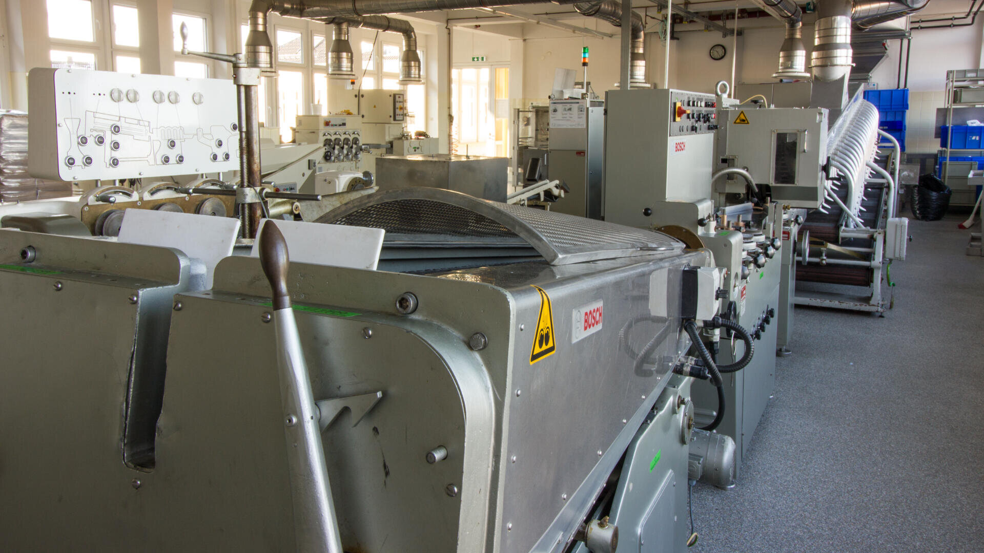 Das Innere einer Fabrik mit mehreren aufgereihten großen Industriemaschinen, ausgestattet mit Bedienfeldern, Warnschildern und Metallkomponenten, in einem gut beleuchteten Raum mit Fenstern und Lüftungsrohren darüber.