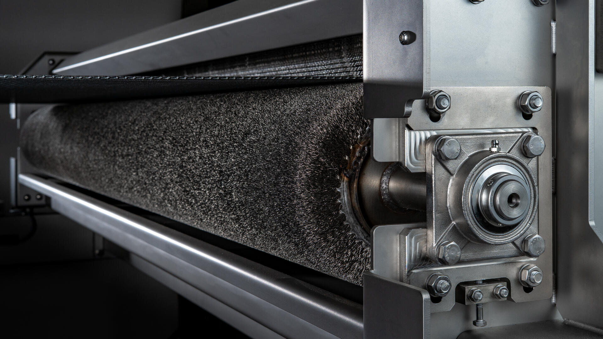 Nahaufnahme einer Industriemaschine mit einer Metallwalzenbürste, die auf einem stabilen Metallrahmen montiert ist, wobei Schrauben und mechanische Komponenten sichtbar sind.
