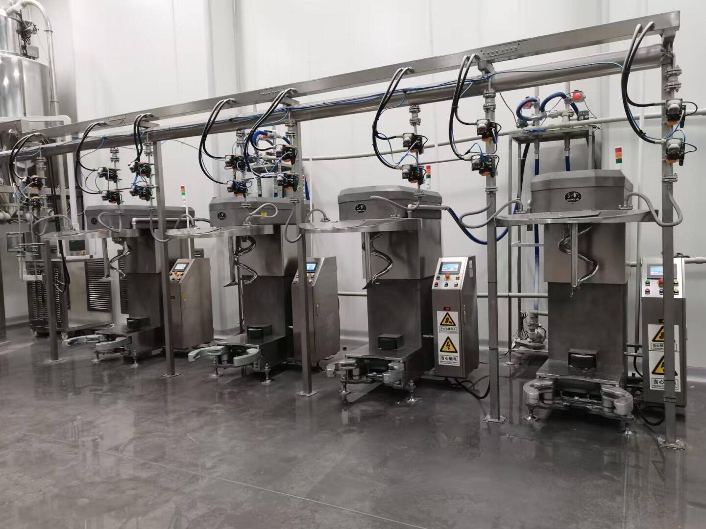 Four stainless steel industrial machines with control panels and warning signs are aligned in a clean, modern factory setting, connected by overhead pipes and cables against a white wall.