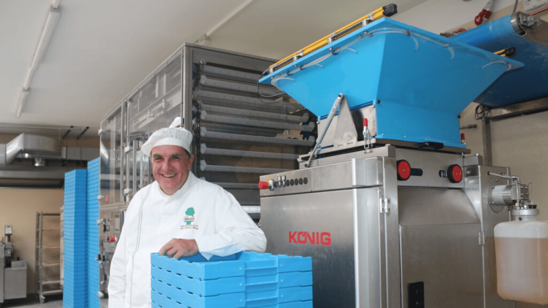 Ein lächelnder Bäcker in weißer Uniform und Hut steht an einer großen industriellen Backmaschine und hält ein blaues Blech in einer hellen, modernen Bäckereiküche.