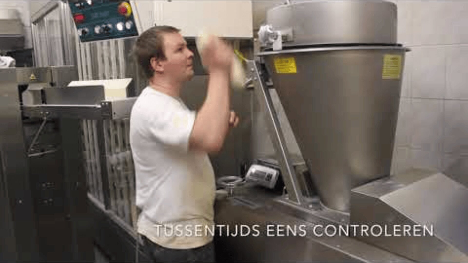 Ein Mann in einem weißen Hemd überprüft eine große Industriemaschine in einer Bäckerei oder Fabrik. Er ist von Schalttafeln und Geräten umgeben. Der niederländische Text am unteren Rand lautet: TUSSENTIJDS EENS CONTROLEREN.