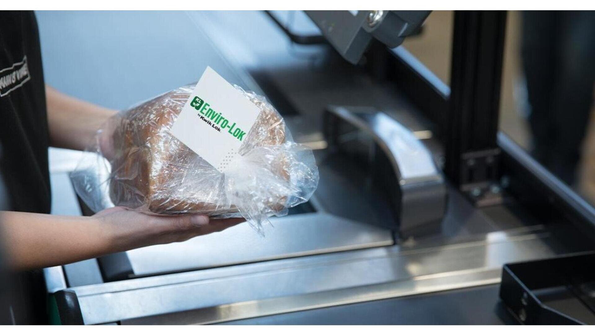 A person holds a loaf of bread wrapped in plastic with an Enviro-Lok label at a checkout counter, ready to scan at a self-service register.