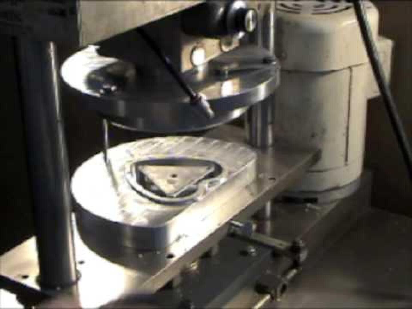 Eine Nahaufnahme einer Metallumformungsmaschine, die ein dreieckiges Stück Metall zwischen zwei runde Matrizen presst. Im Hintergrund sind mechanische Komponenten und ein Motor zu sehen.