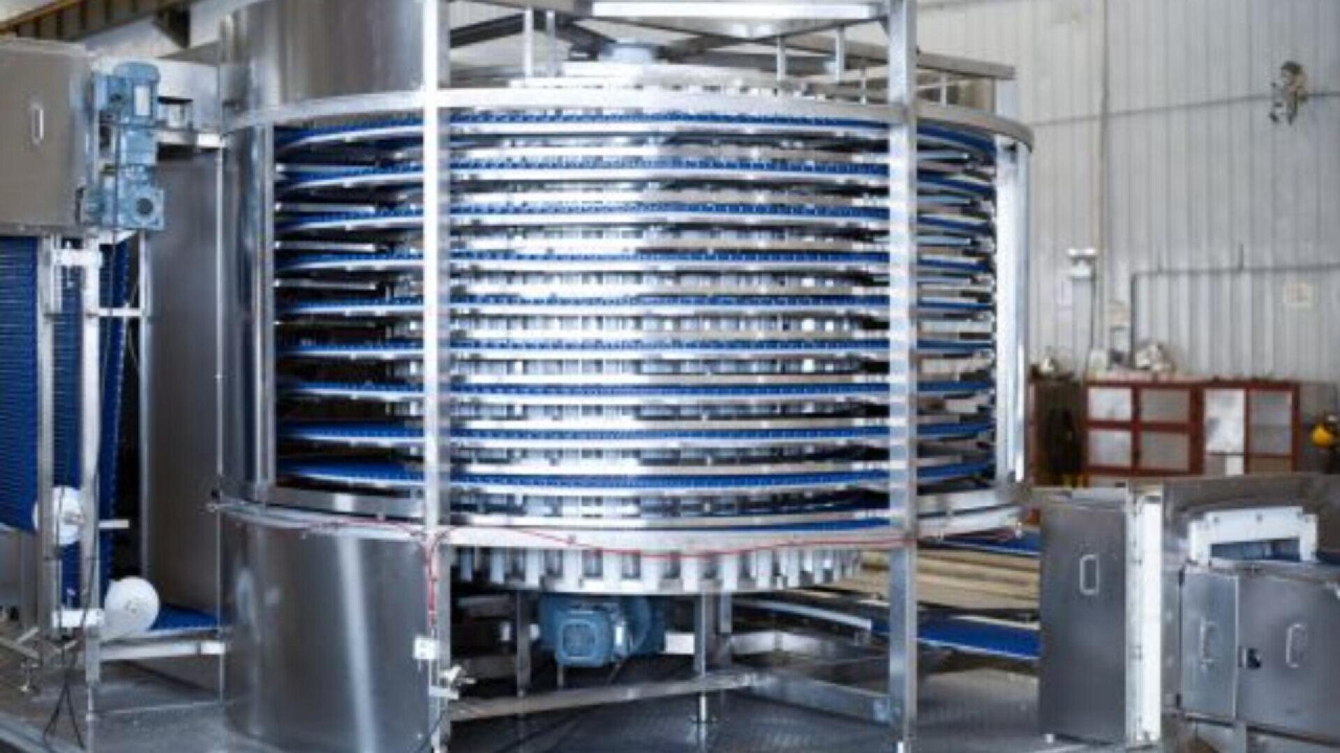 Ein großes industrielles Spiralfördersystem aus Metall mit mehreren Etagen und blauen Förderbändern befindet sich in einer Fabrik oder einem Verarbeitungsbetrieb.