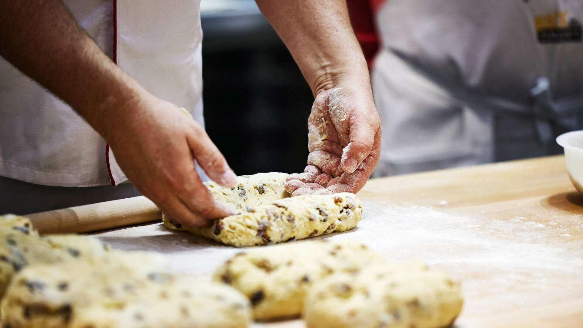 Eine Person formt auf einer bemehlten Holzfläche einen Teig mit Rosinen oder Schokoladenstückchen und bereitet ihn zum Backen vor. Weitere Teigstücke und ein Nudelholz sind in der Nähe zu sehen.