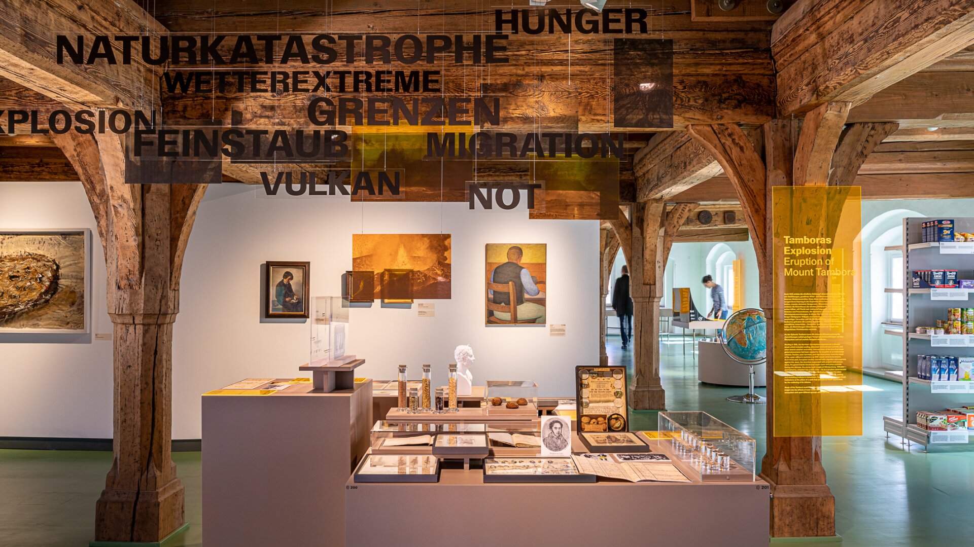 Eine Museumsausstellung zeigt Artefakte, Kunstwerke und historische Gegenstände unter Holzbalken. Über den Exponaten schweben deutsche Wörter wie Naturkatastrophe, Explosion und Migration.