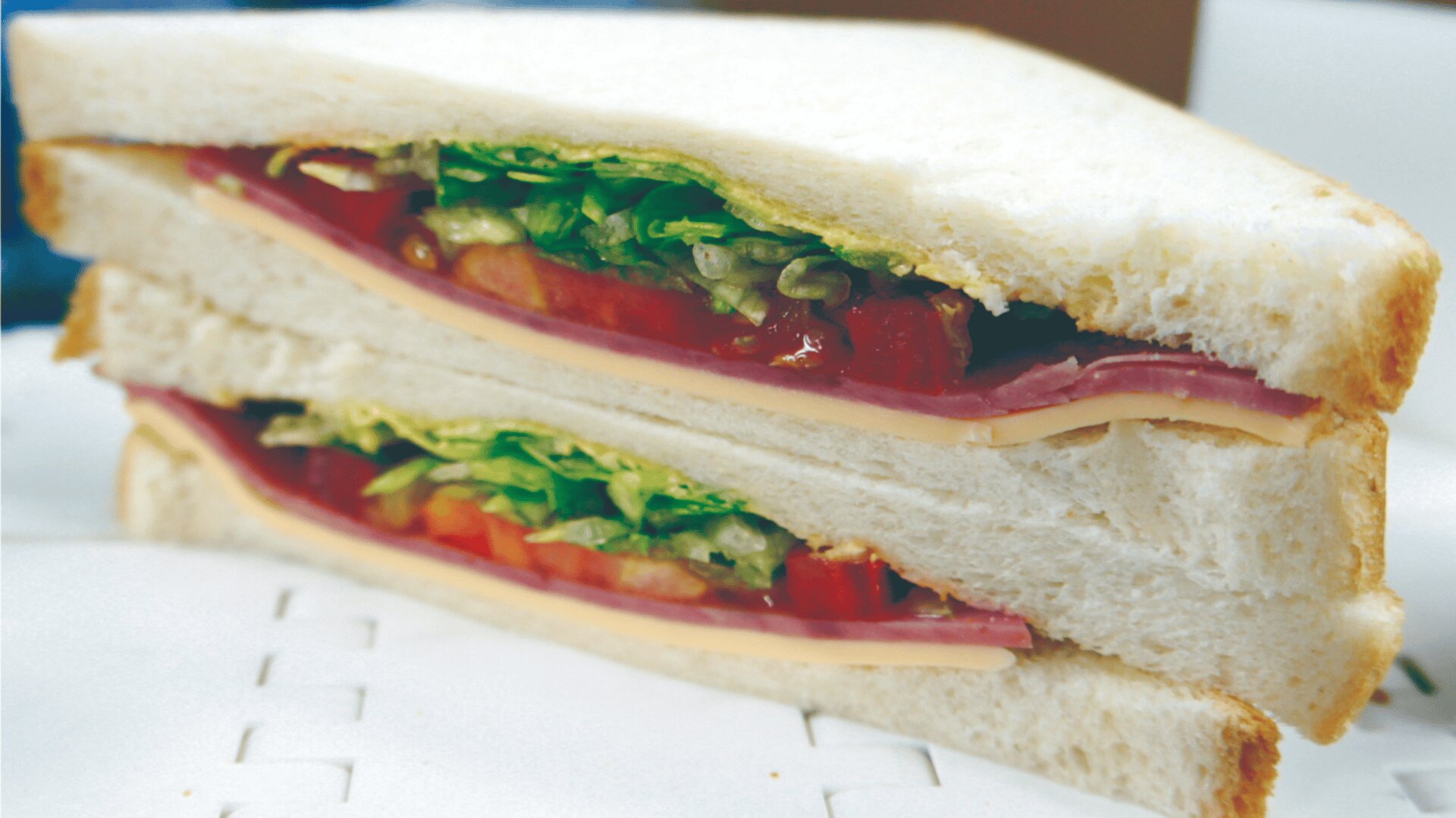 Nahaufnahme eines halbierten Sandwichs mit Schichten von Weißbrot, Schinken, Käse, Salat und Tomate. Die Sandwichhälften werden gestapelt und auf eine weiße Fläche gelegt.
